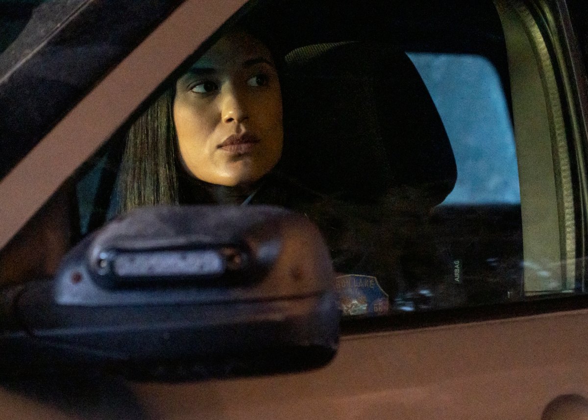 Julia Jones as Angela in Dexter: New Blood. Angela sits in her police car and looks out the window.