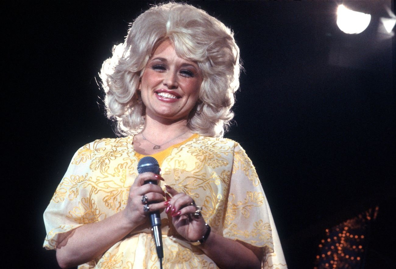 Dolly Parton wears a yellow dress and holds a microphone. 