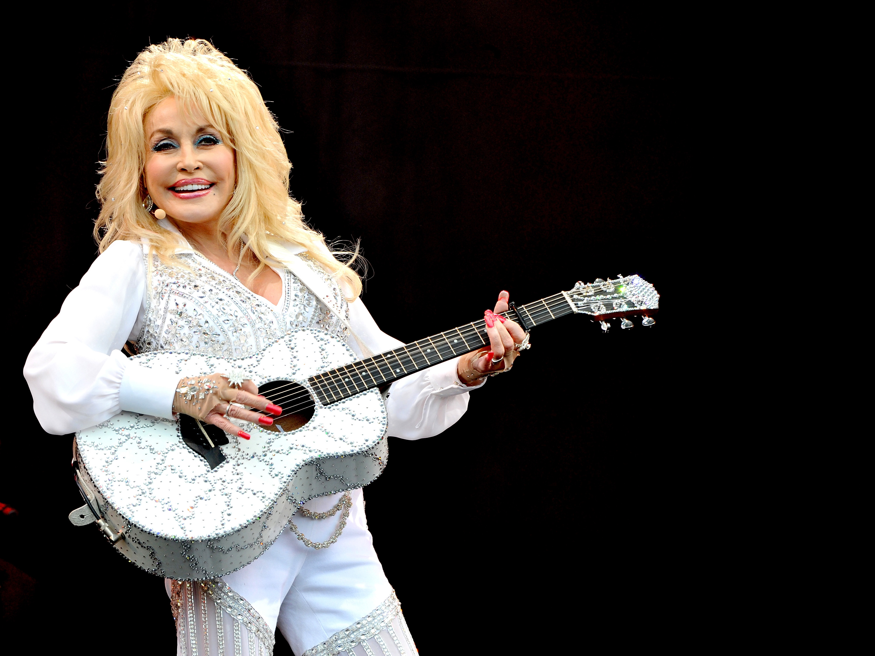 Dolly Parton wears a white rhinestoned outfit and holds a white rhinestoned guitar.