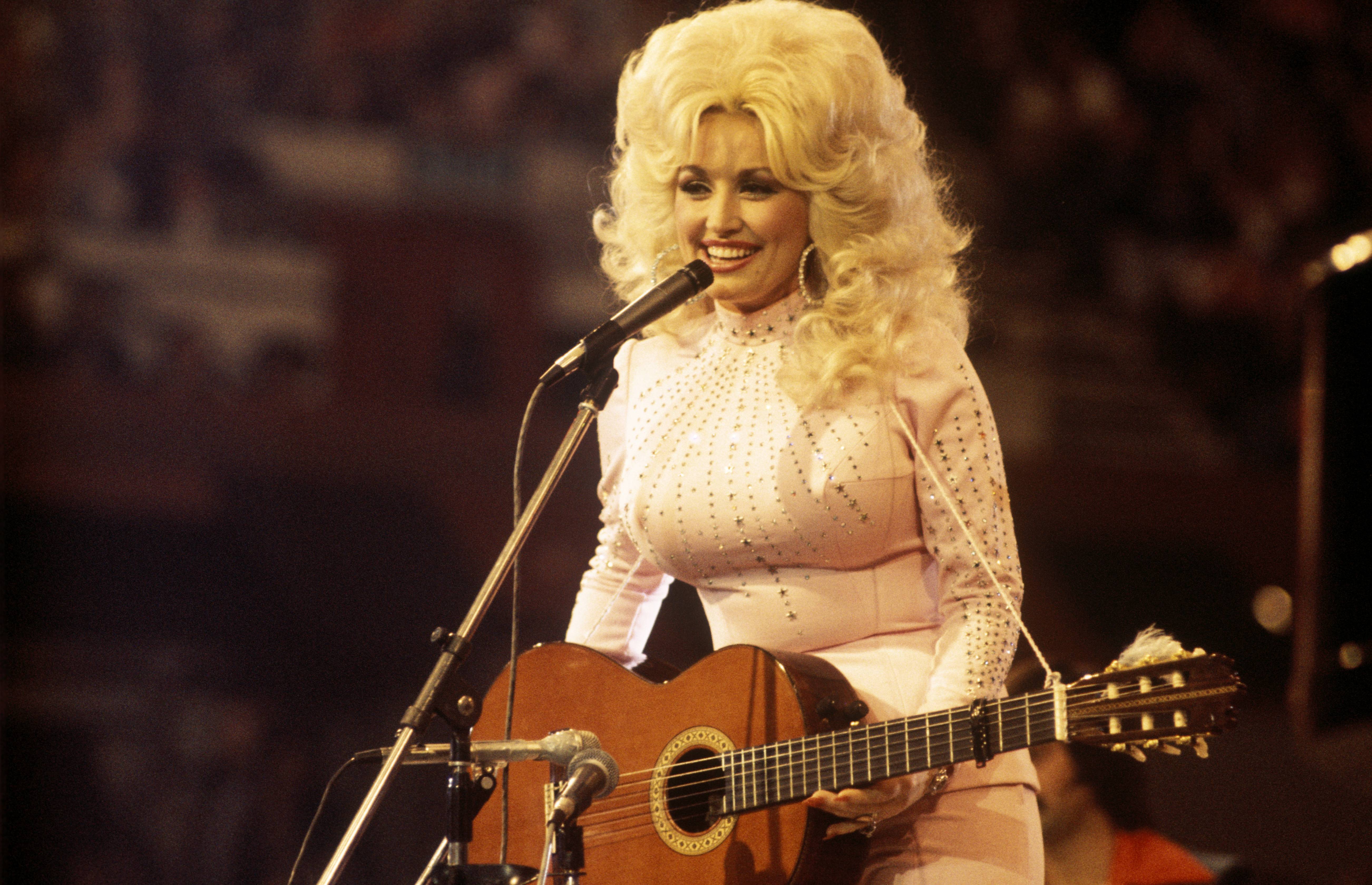 Dolly Parton wears a white turtleneck and holds a guitar. She stands in front of a microphone. 