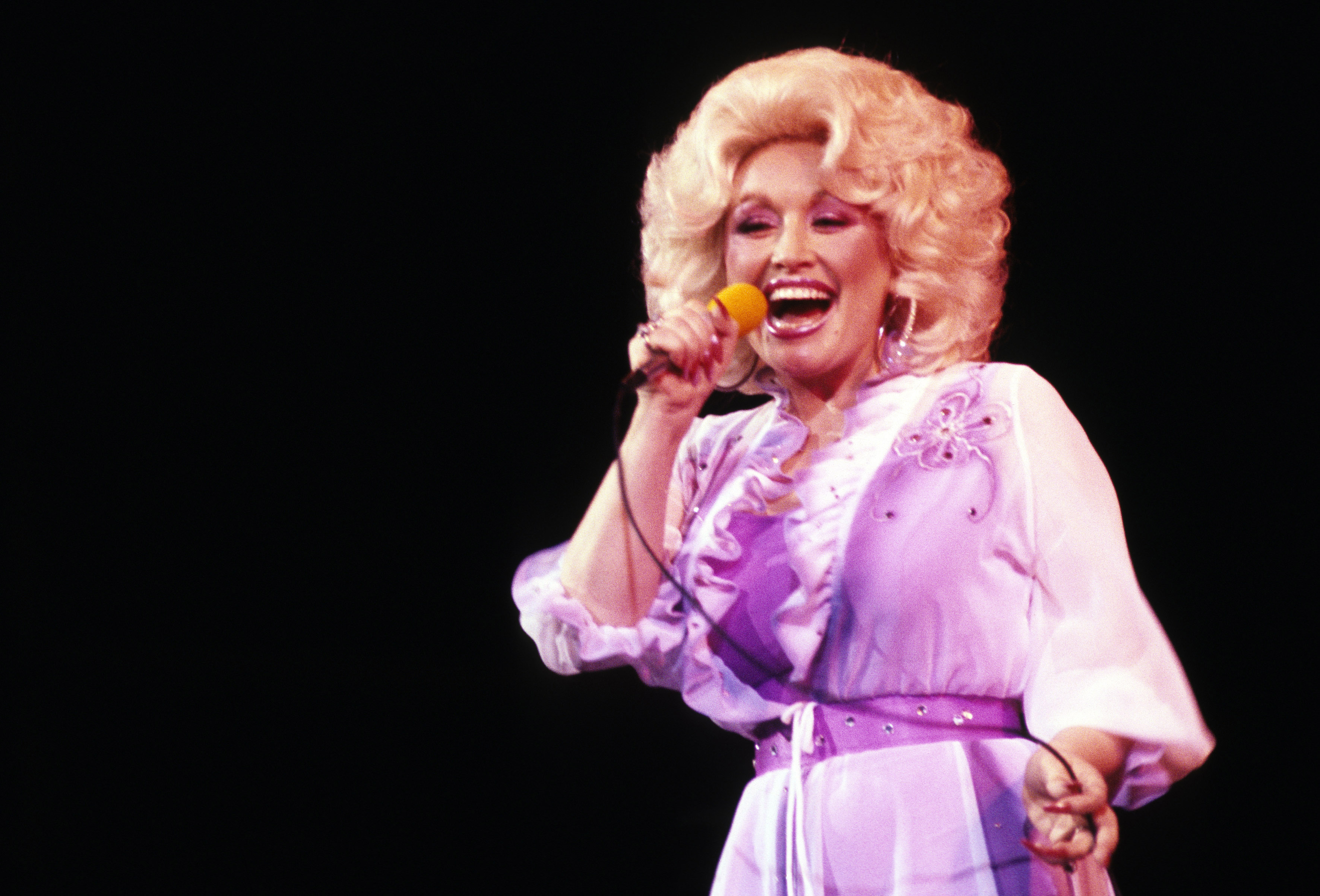Dolly Parton wears a purple dress and sings into a microphone.