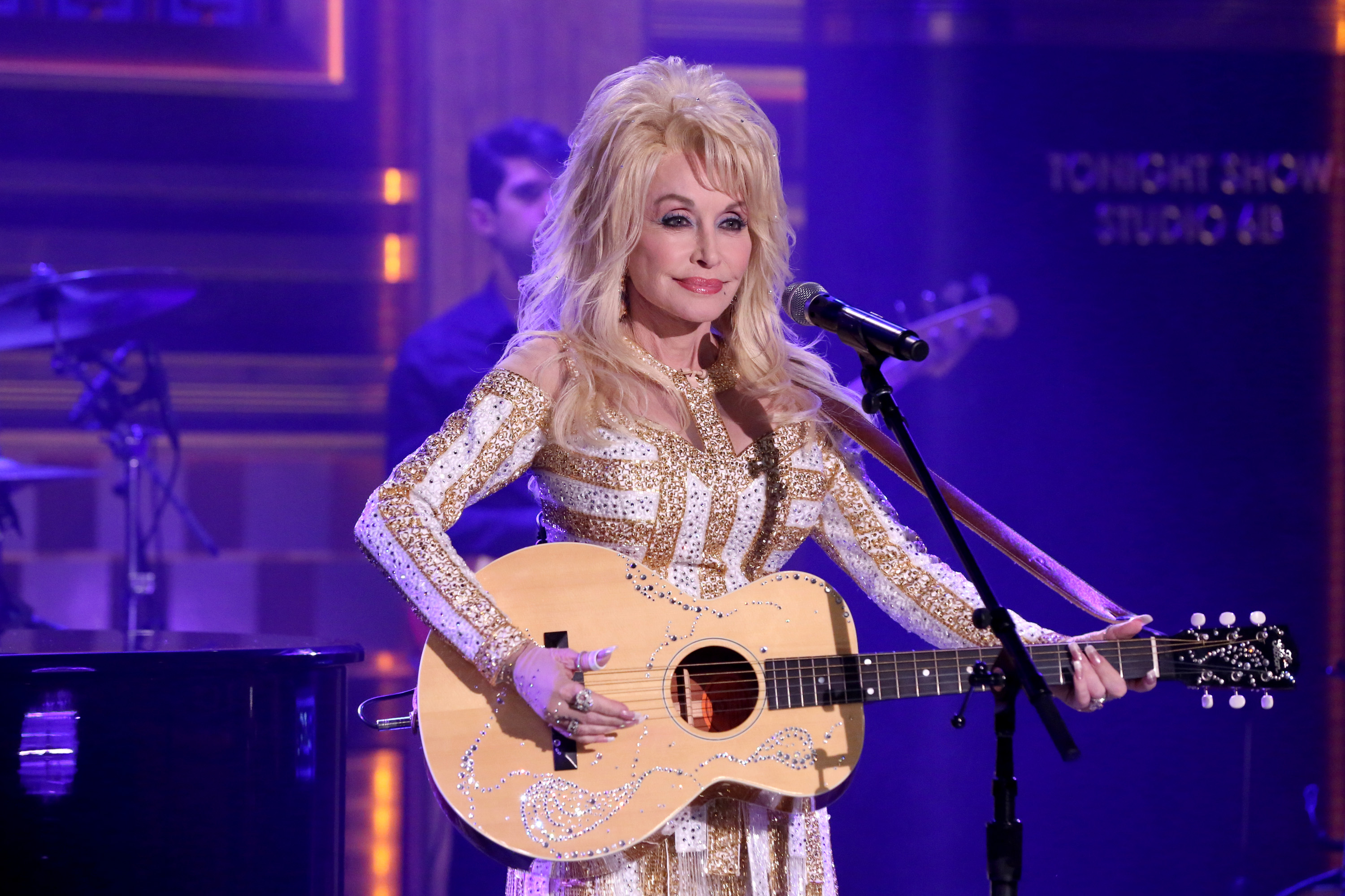 Dolly Parton wears a gold and white shirt and holds a guitar. She stands in front of a microphone.