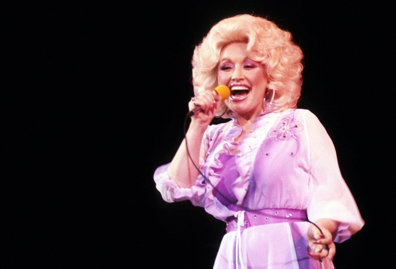 Dolly Parton wears a purple dress and holds a microphone. Dolly Parton's husband tends to stay out of the spotlight.