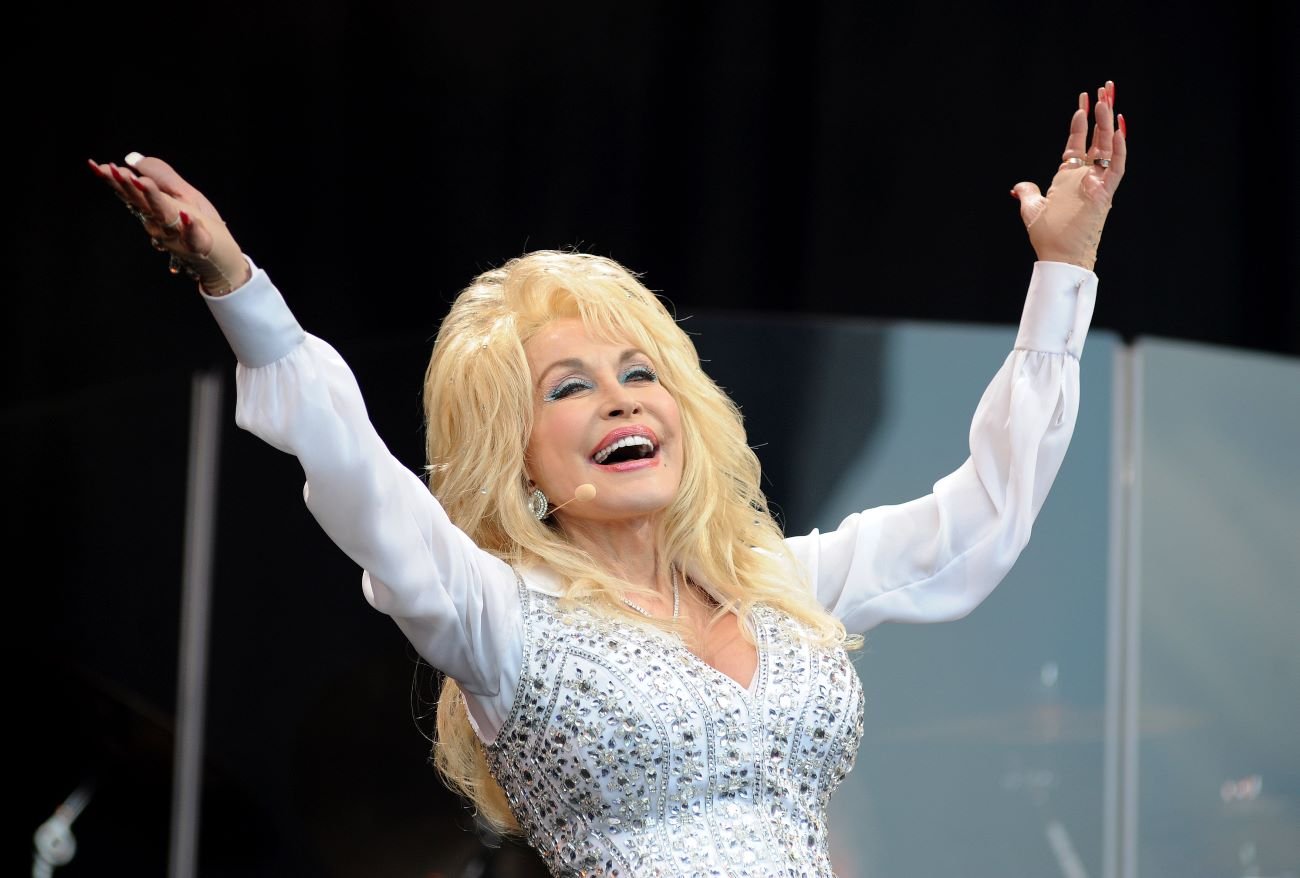 Dolly Parton wears a white, bedazzled shirt and holds her hands up above her head while on stage.
