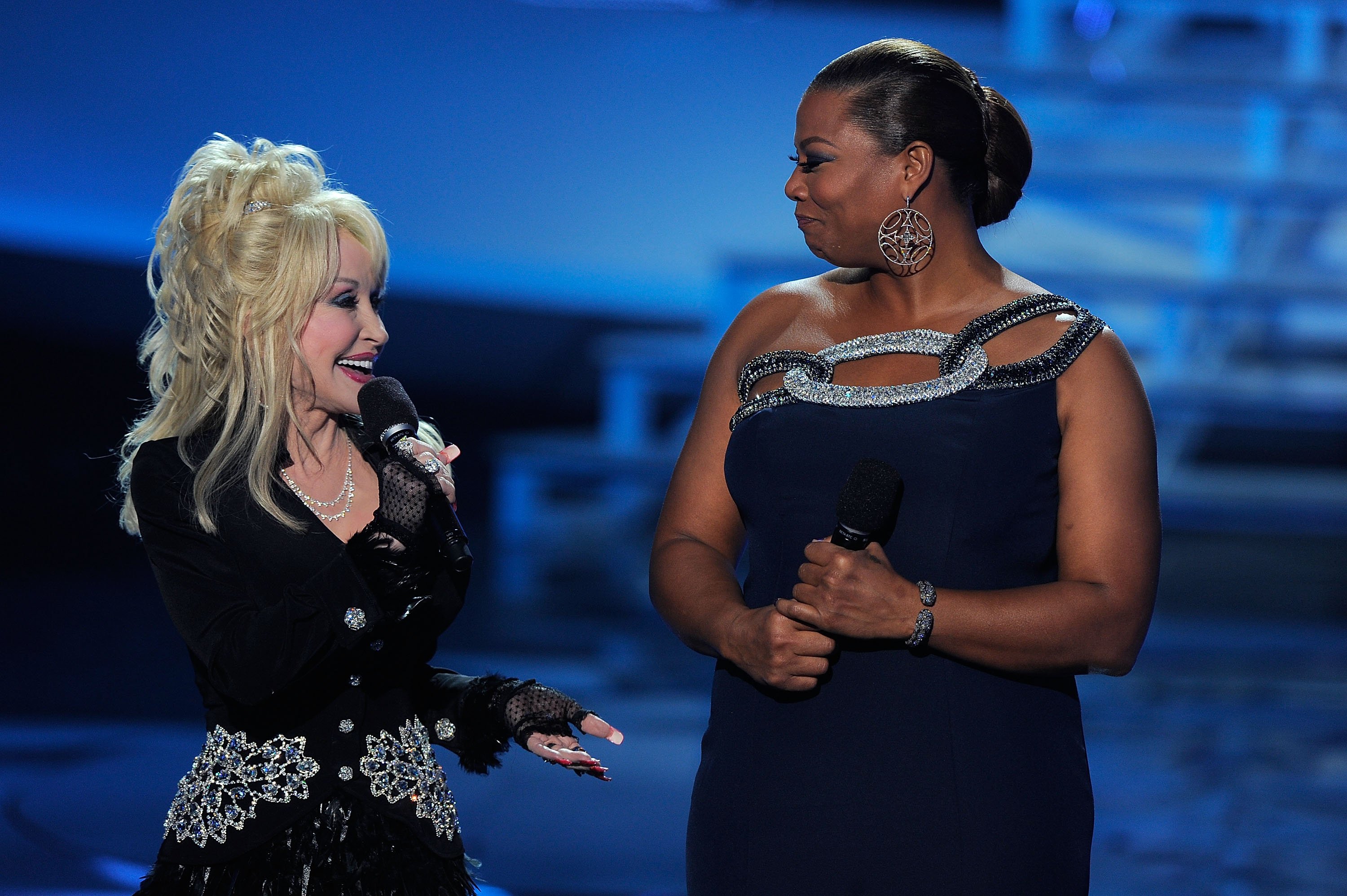 Dolly Parton holds a microphone and smiles while looking at Queen Latifah.