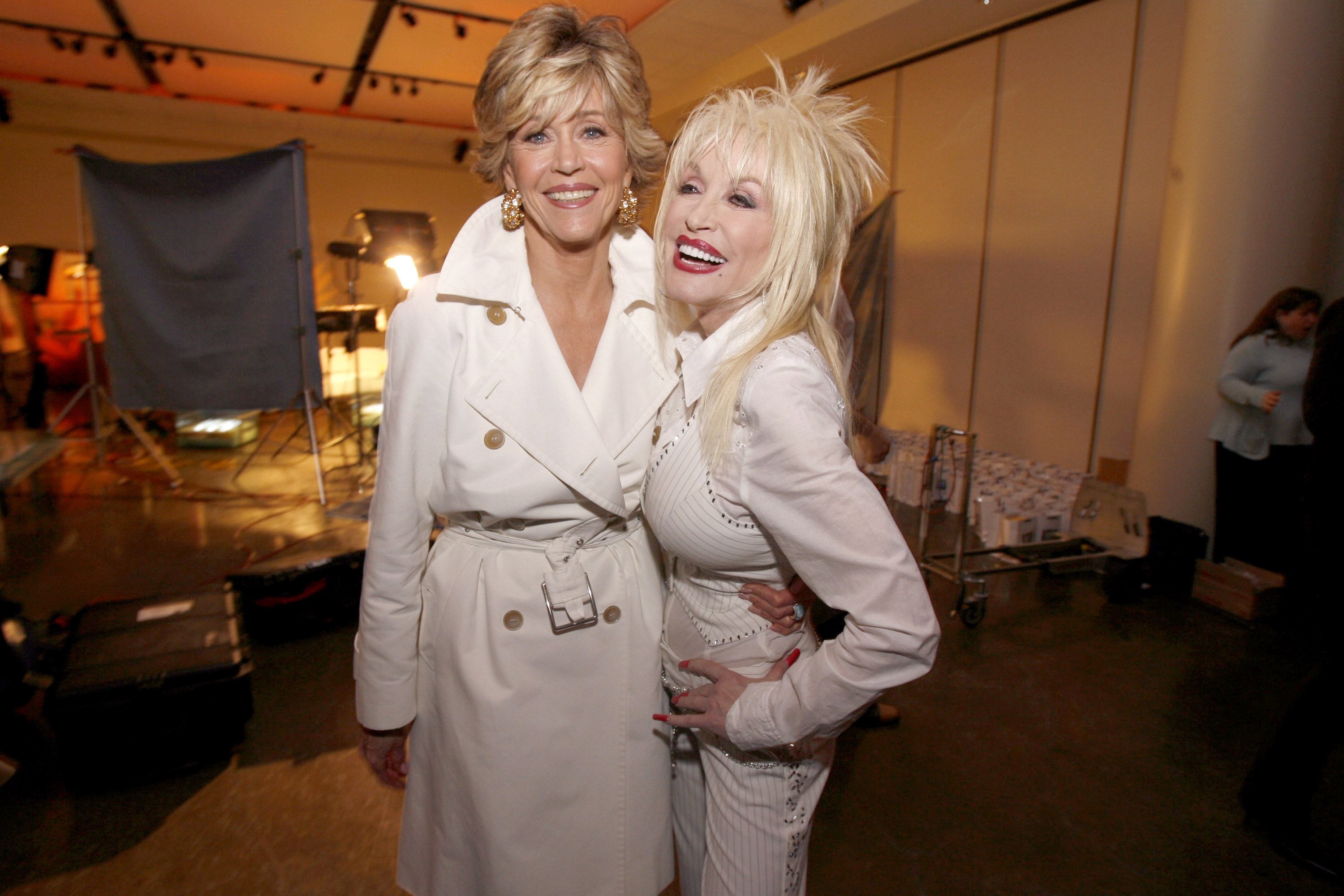 9 to 5 costars Jane Fonda and Dolly Parton wear white outfits and stand with their arms around each other.