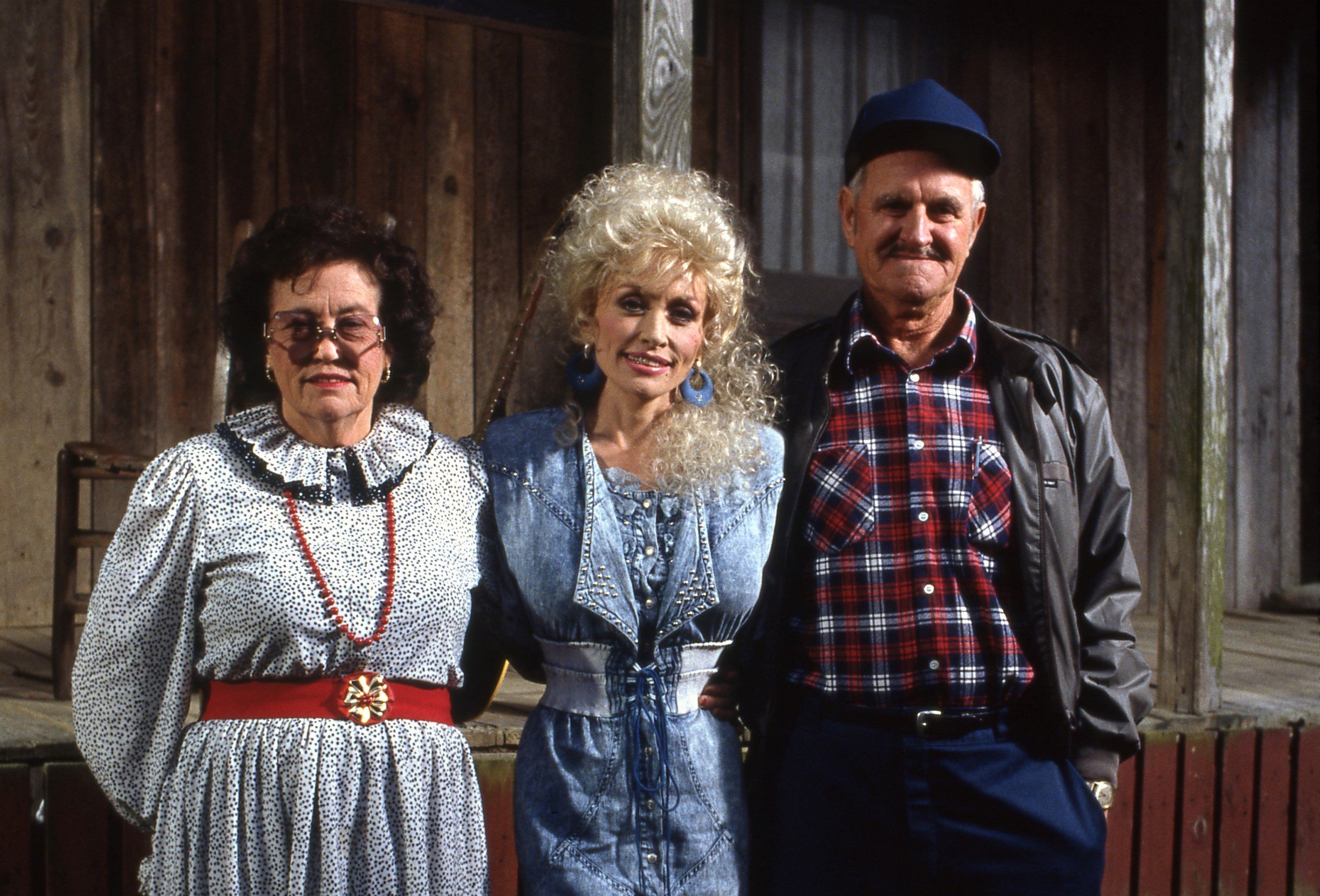 Dolly Parton stands in between her parents Avie Lee Owens and Lee Parton. They stand in front of a log cabin. Dolly Parton has eleven siblings.
