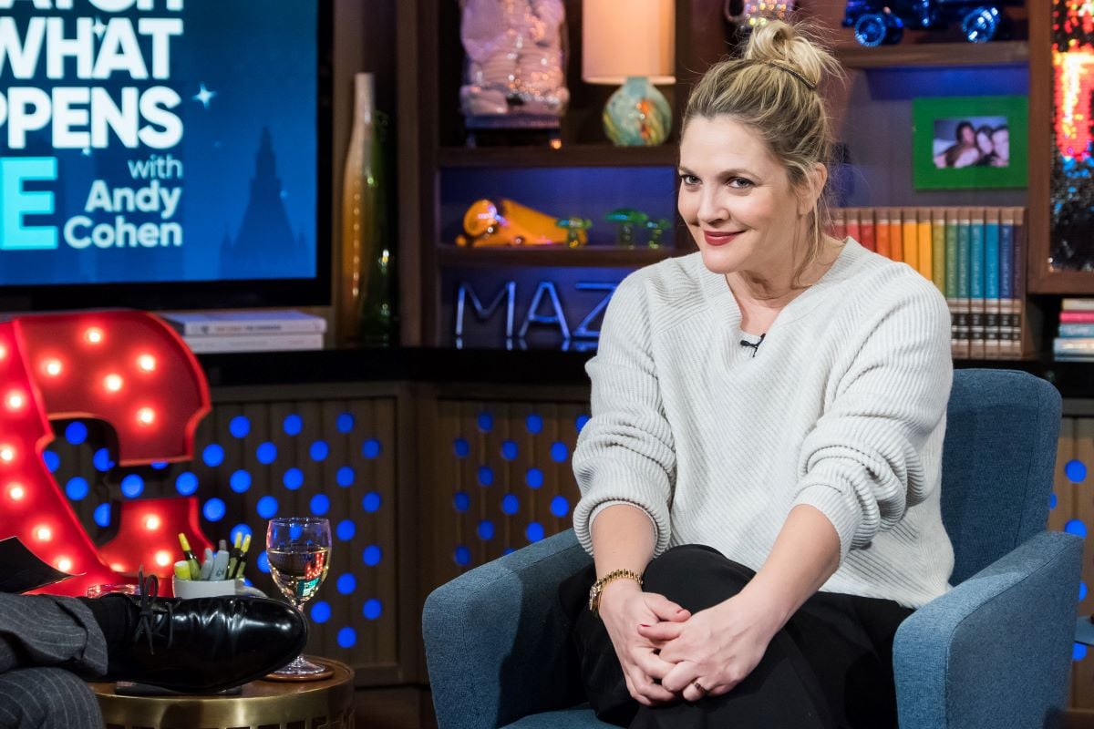 Drew Barrymore seated with hair in a bun, wearing an off-white sweater and black pants