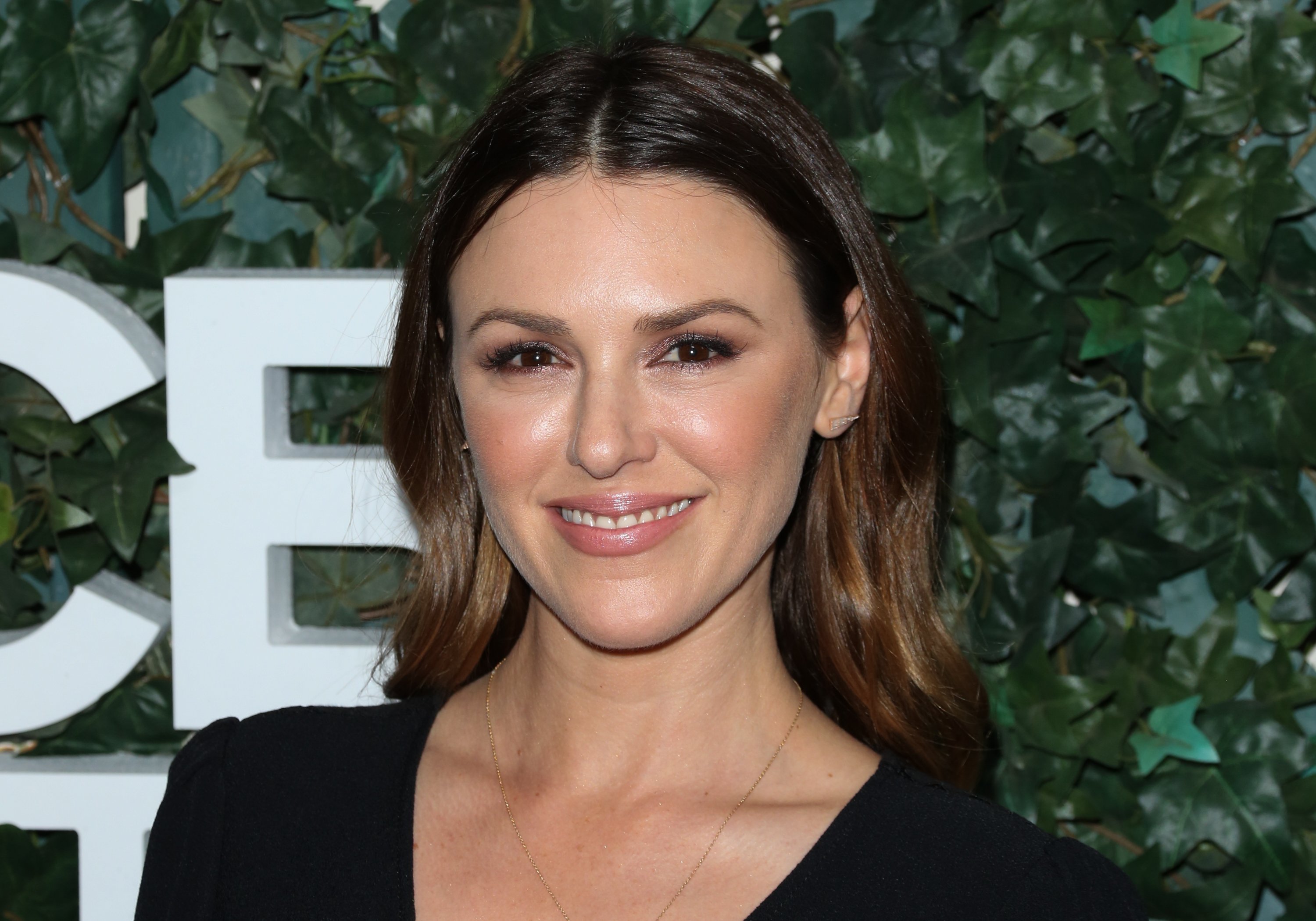 'The Young and the Restless' actor Elizabeth Hendrickson wears a black jumpsuit and poses in front of a hedge.