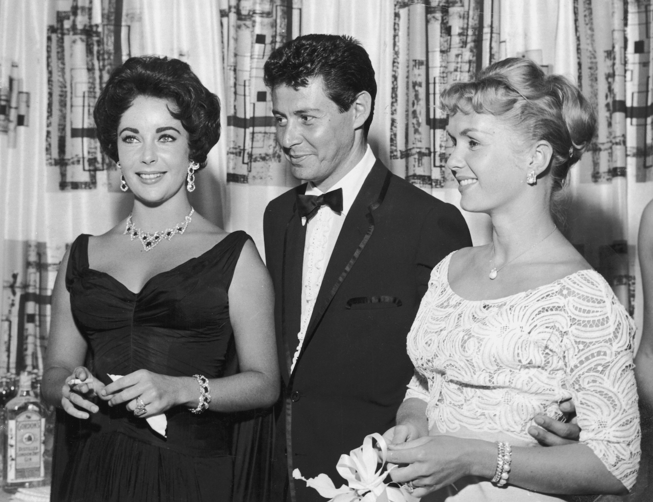 Eddie Fisher, wearing a tuxedo, stands with arm around his wife, American actor Debbie Reynolds (R) and smiles while looking at British-born actor Elizabeth Taylor, smoking a cigarette in 1958.