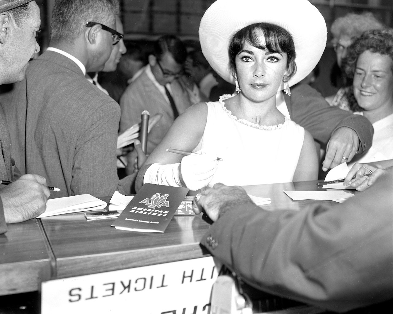 Elizabeth Taylor in a white dress and hat, standing with a pen in her hand