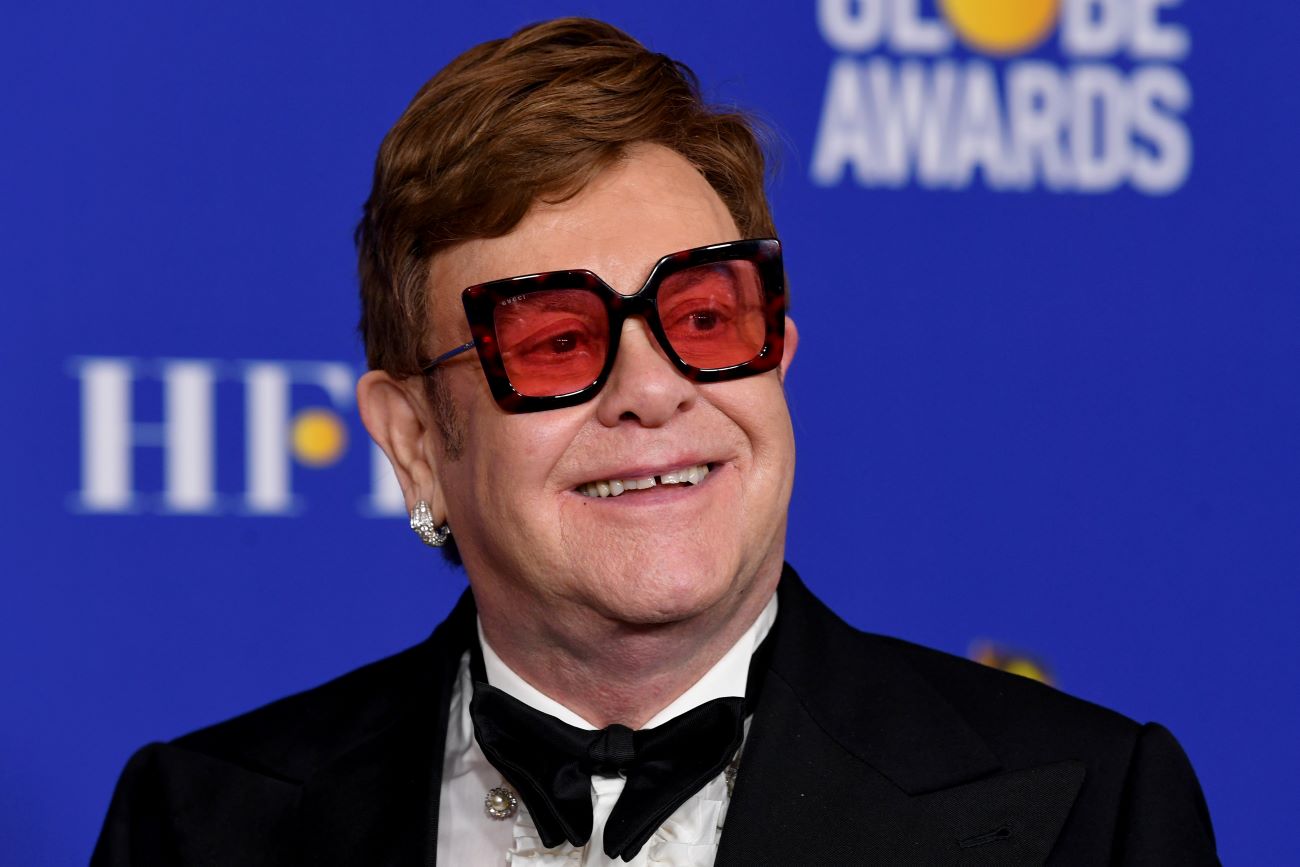 Elton John wears a tuxedo and sunglasses and stands in front of a blue background.