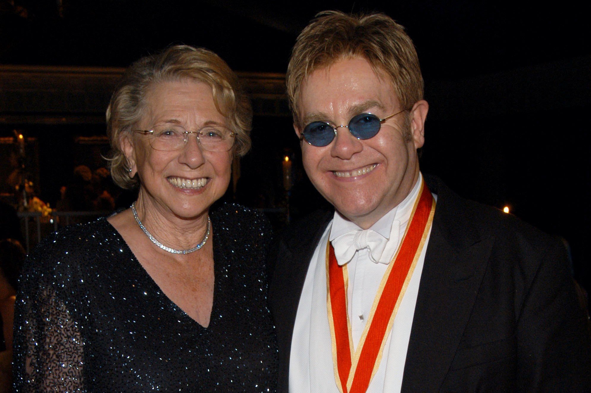 Elton John and his mother stand next to each other and smile. He wears sunglasses.