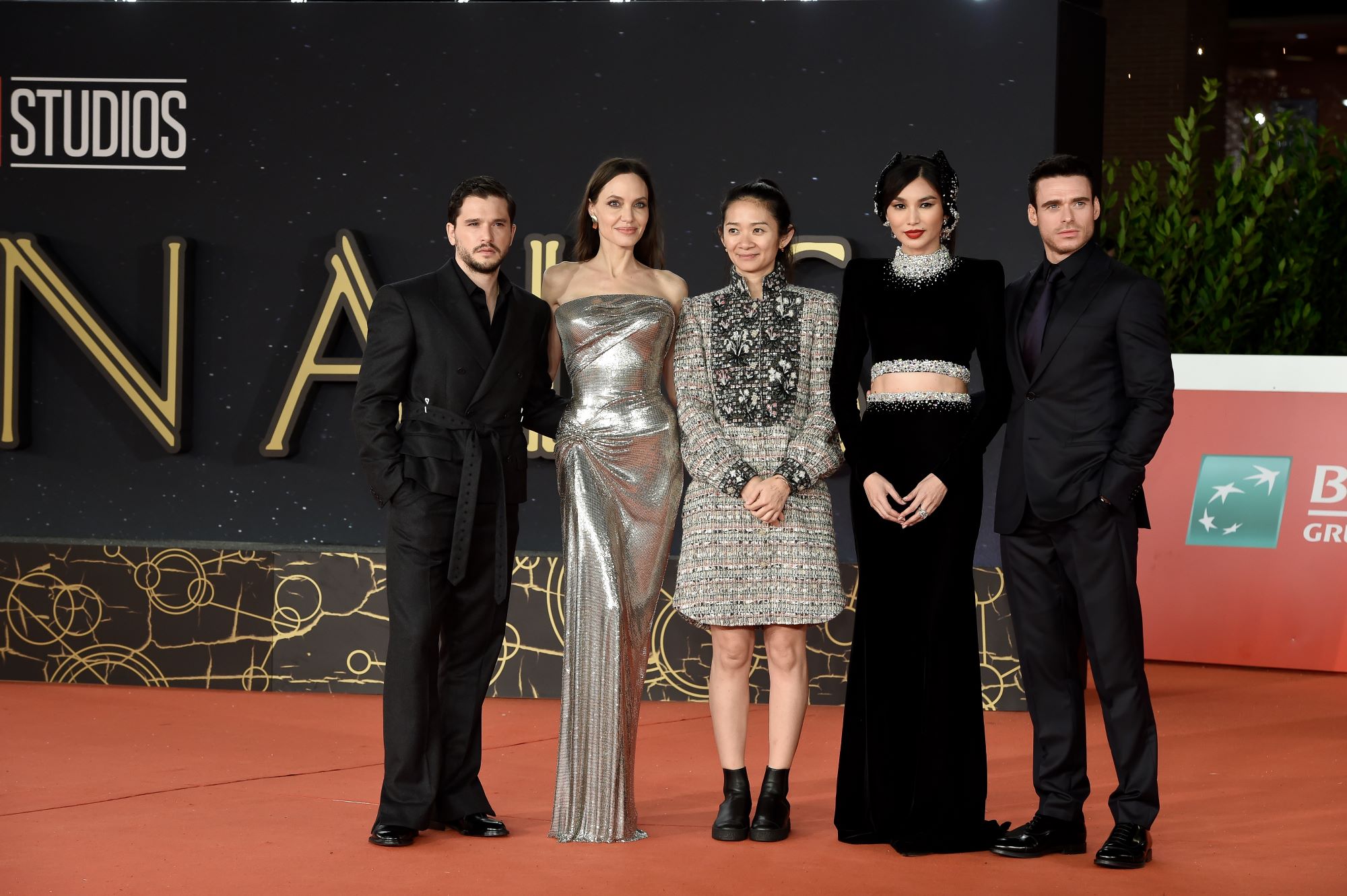 The 'Eternals' cast and crew, Kit Harington, Angelina Jolie, Chloé Zhao, Gemma Chan, and Richard Madden, pose together on the red carpet. Harington and Madden wears black suits. Jolie wears a long strapless silver dress. Zhao wears a knee-length gray and black long-sleeved dress. Chan wears a long black long-sleeved dress. 'Eternals' is now on Disney+ and is rumored to have a sequel in the works.