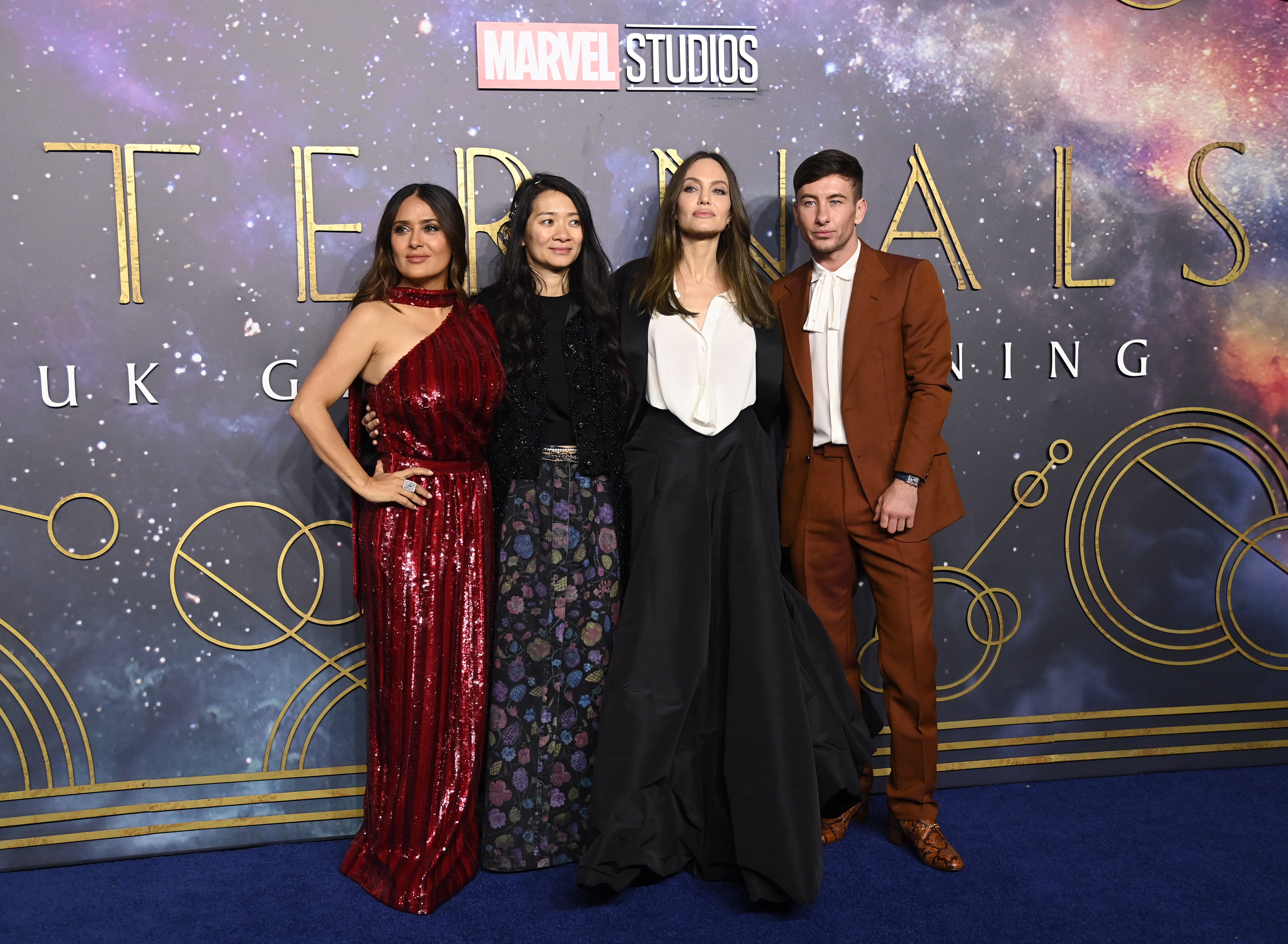 Salma Hayek, Chloe Zhao, Angelina Jolie, and Barry Keoghan pose for pictures on the red carpet at the 'Eternals' premiere. Hayek wears a sparkly red dress. Zhao wears a black top and long floral skirt. Jolie, who is in an 'Eternals' post-credits scene, wears a black and white dress. Keoghan wears a brown suit over a white shirt.