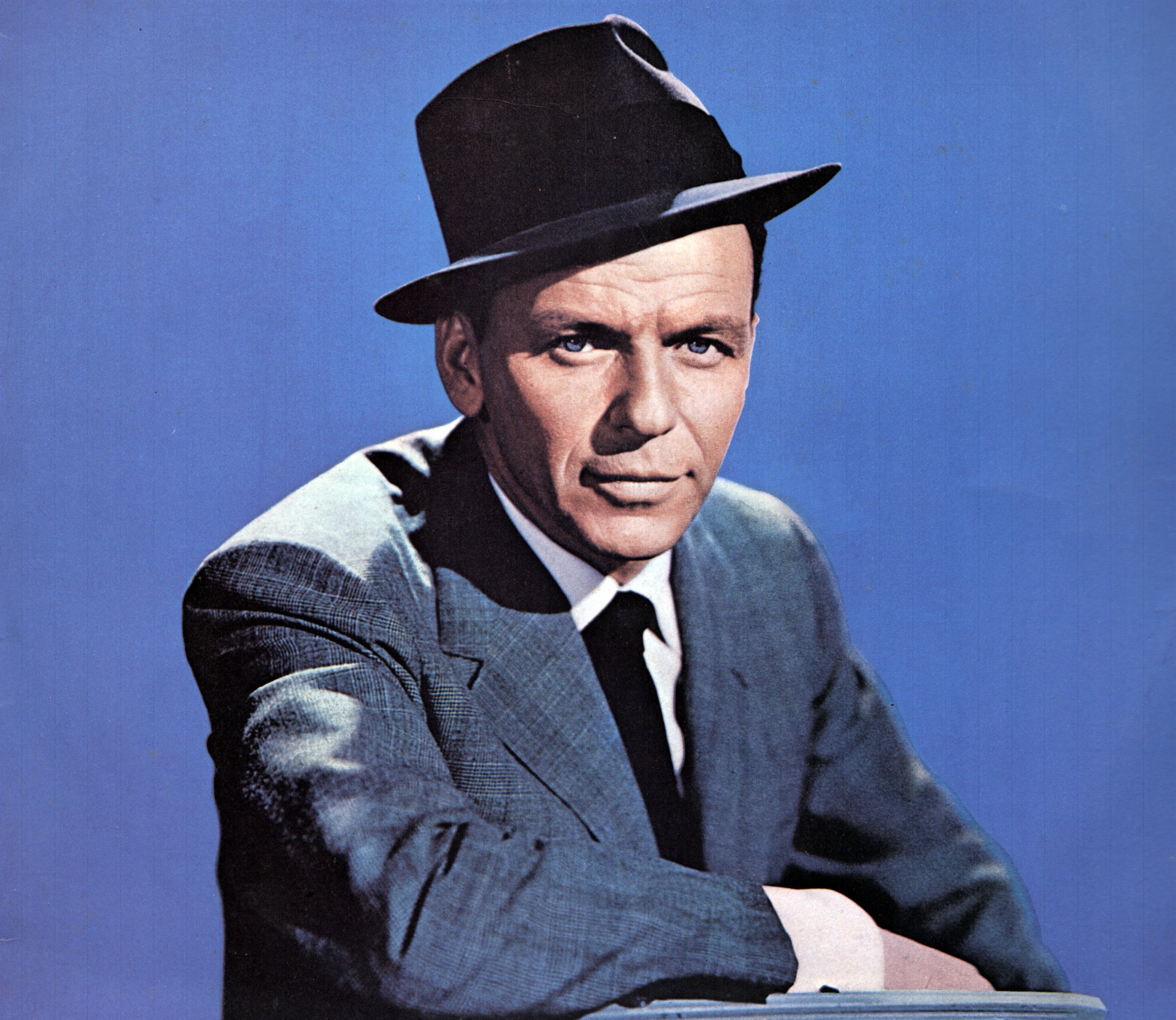 Frank Sinatra wears a suit and hat and sits against a blue background.