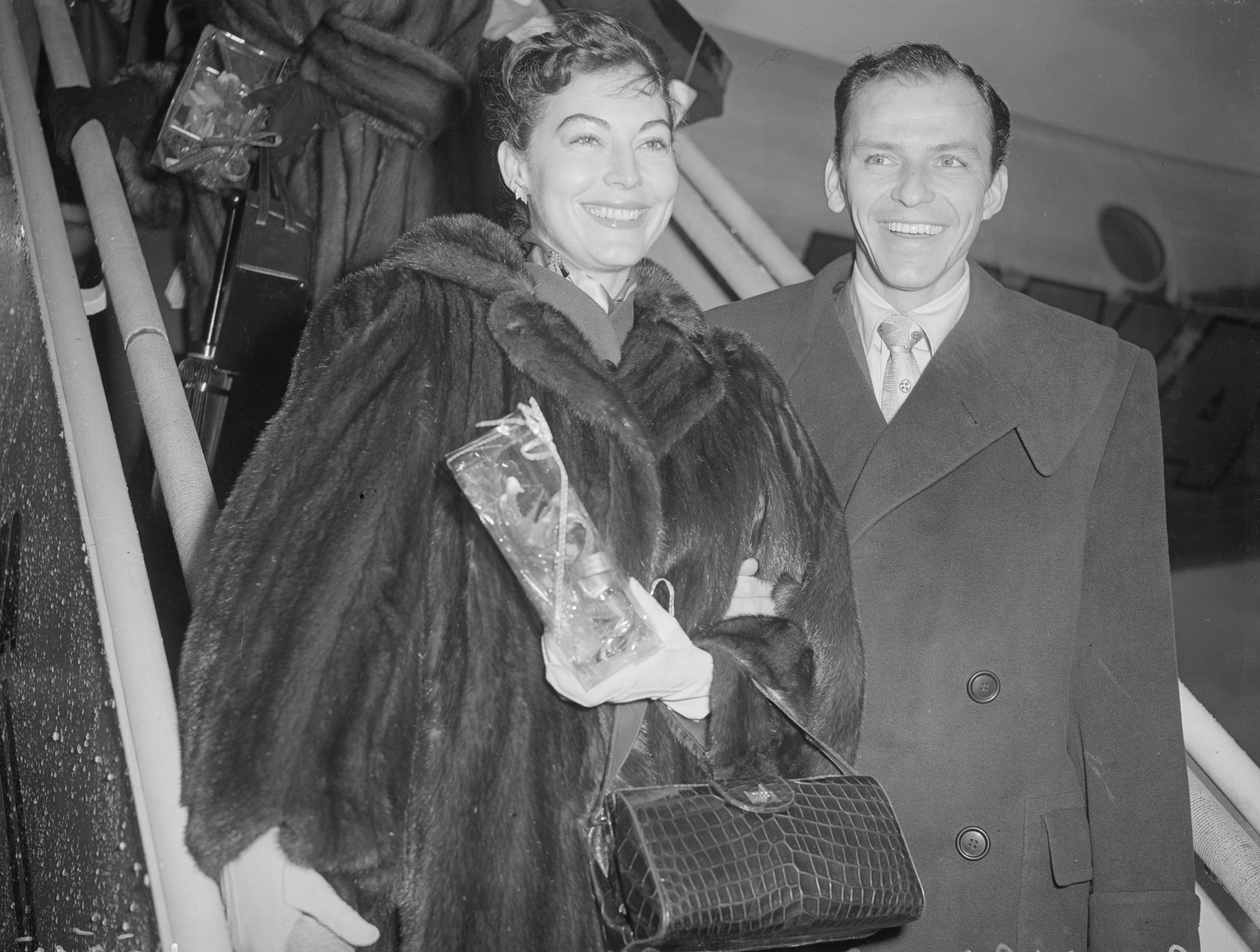 Ava Gardner wears a fur coat and Frank Sinatra wears a coat and tie. They descend the steps from an airplane.