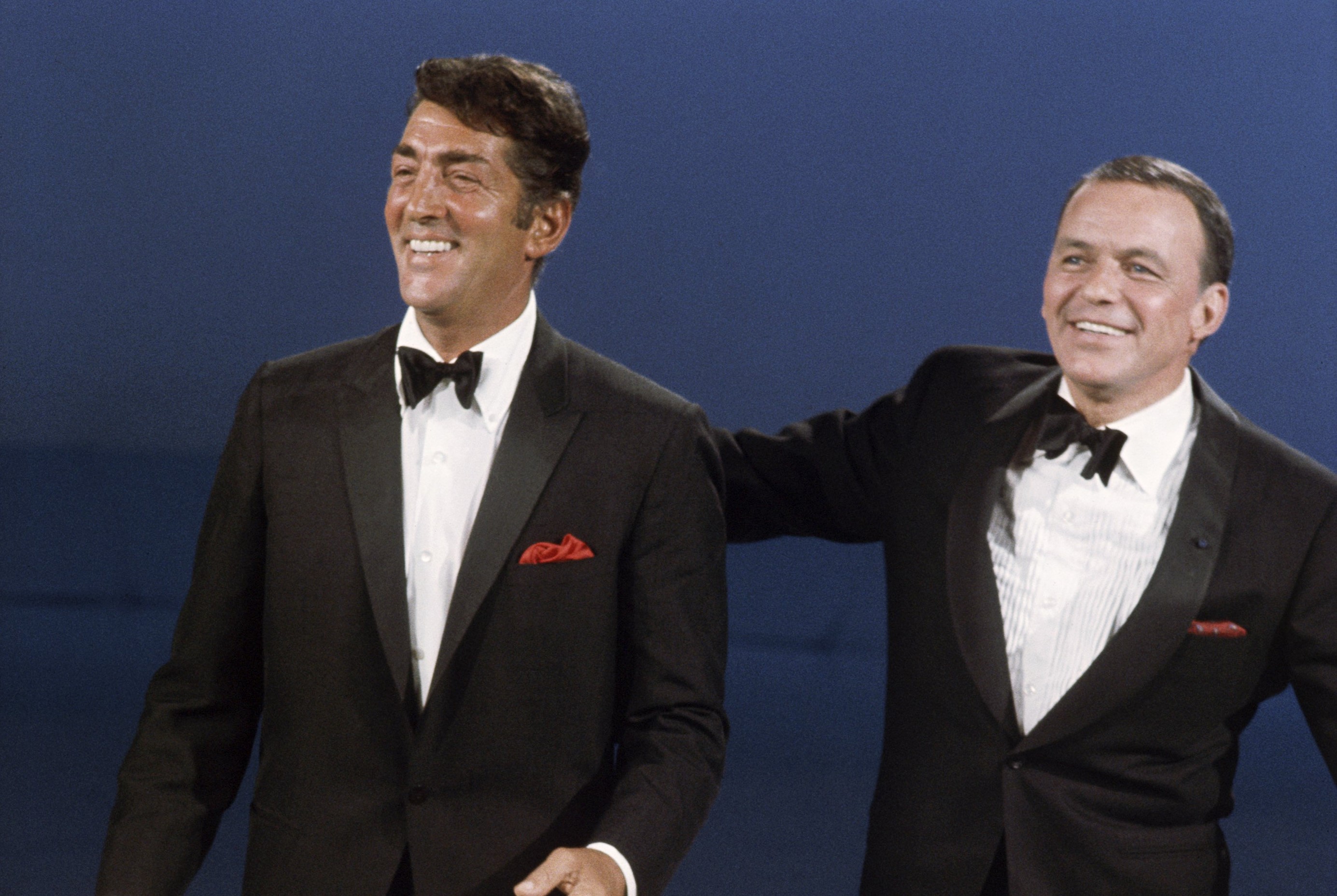 Frank Sinatra and Dean Martin wear tuxedos with red pocket squares. Sinatra stands with his hand on Martin's back.