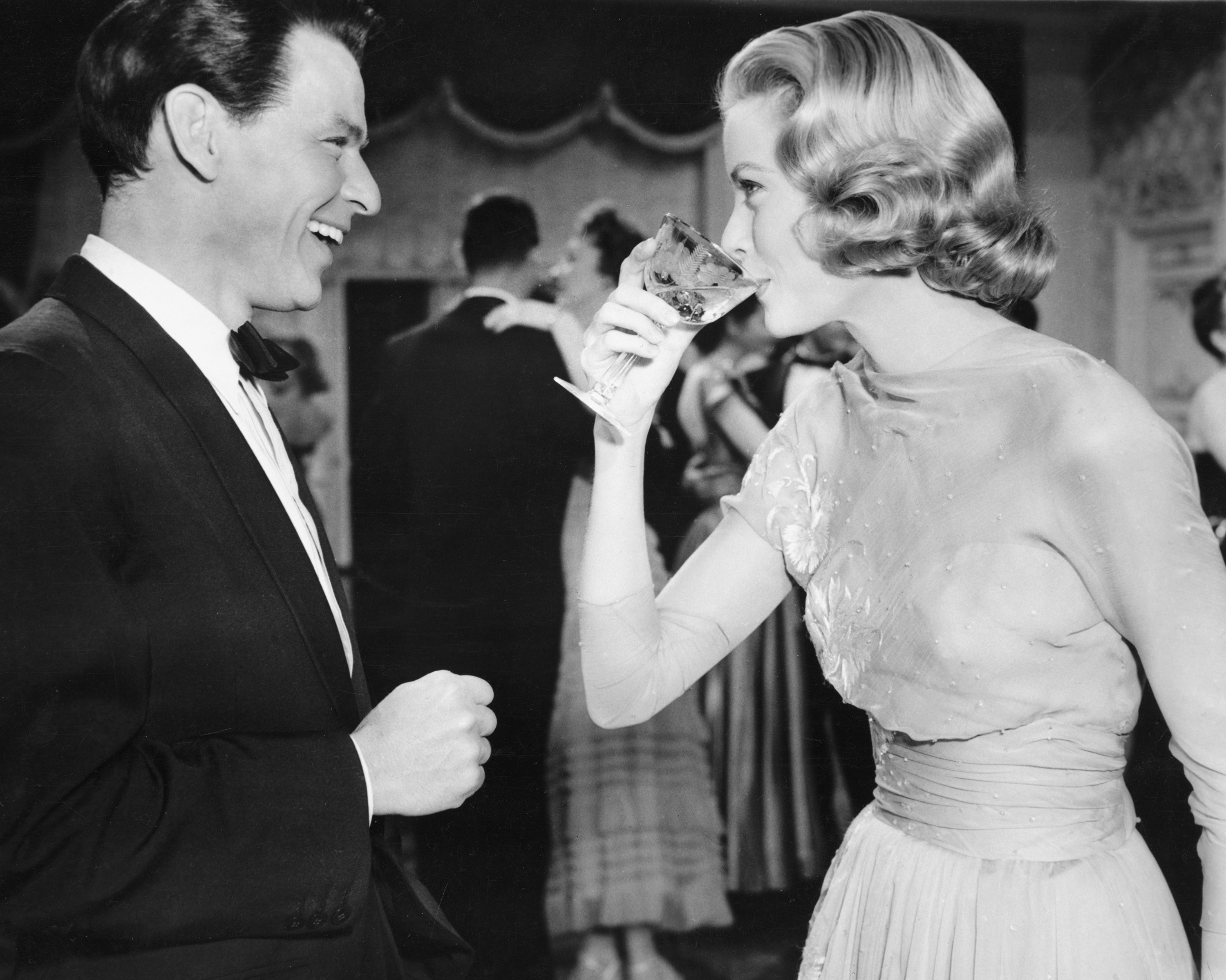 Frank Sinatra wears a tuxedo and smiles at Grace Kelly, who drinks from a glass.