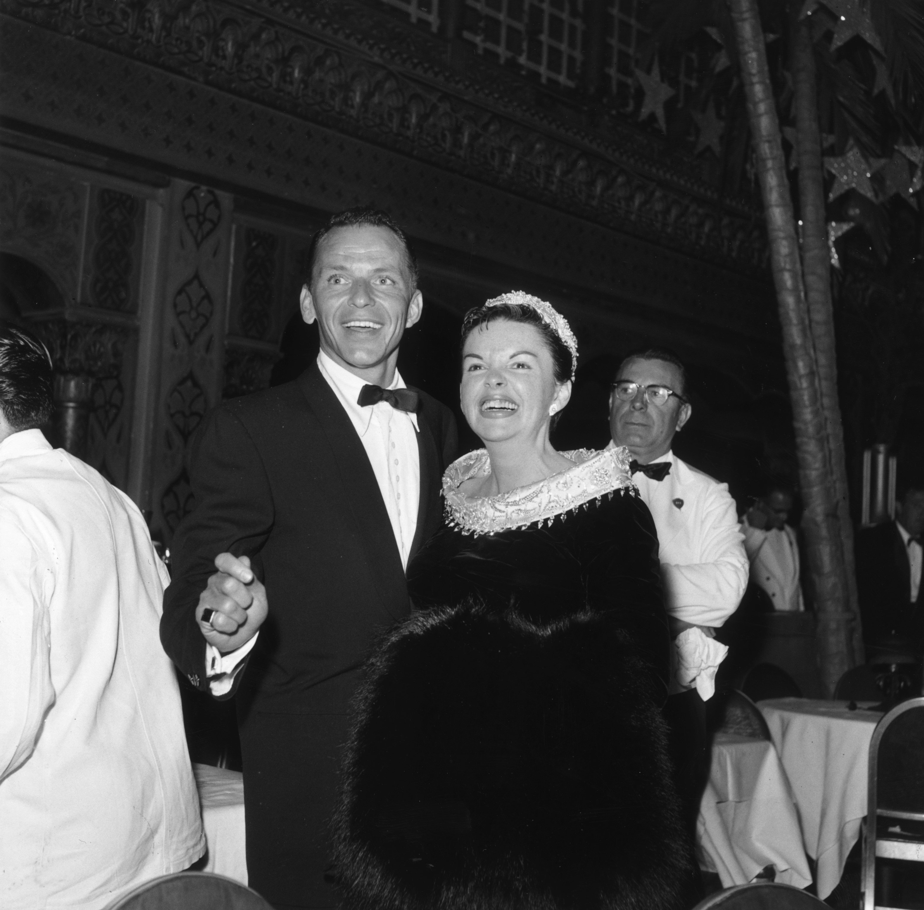 Frank Sinatra wears a suit and Judy Garland wears a black dress with a white collar. They stand together and smile.
