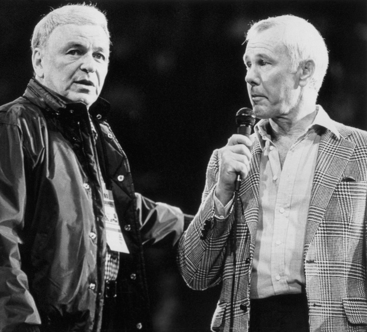 Frank Sinatra in a windbreaker and standing beside Johnny Carson, whose in a plaid jacket and holding a microphone.