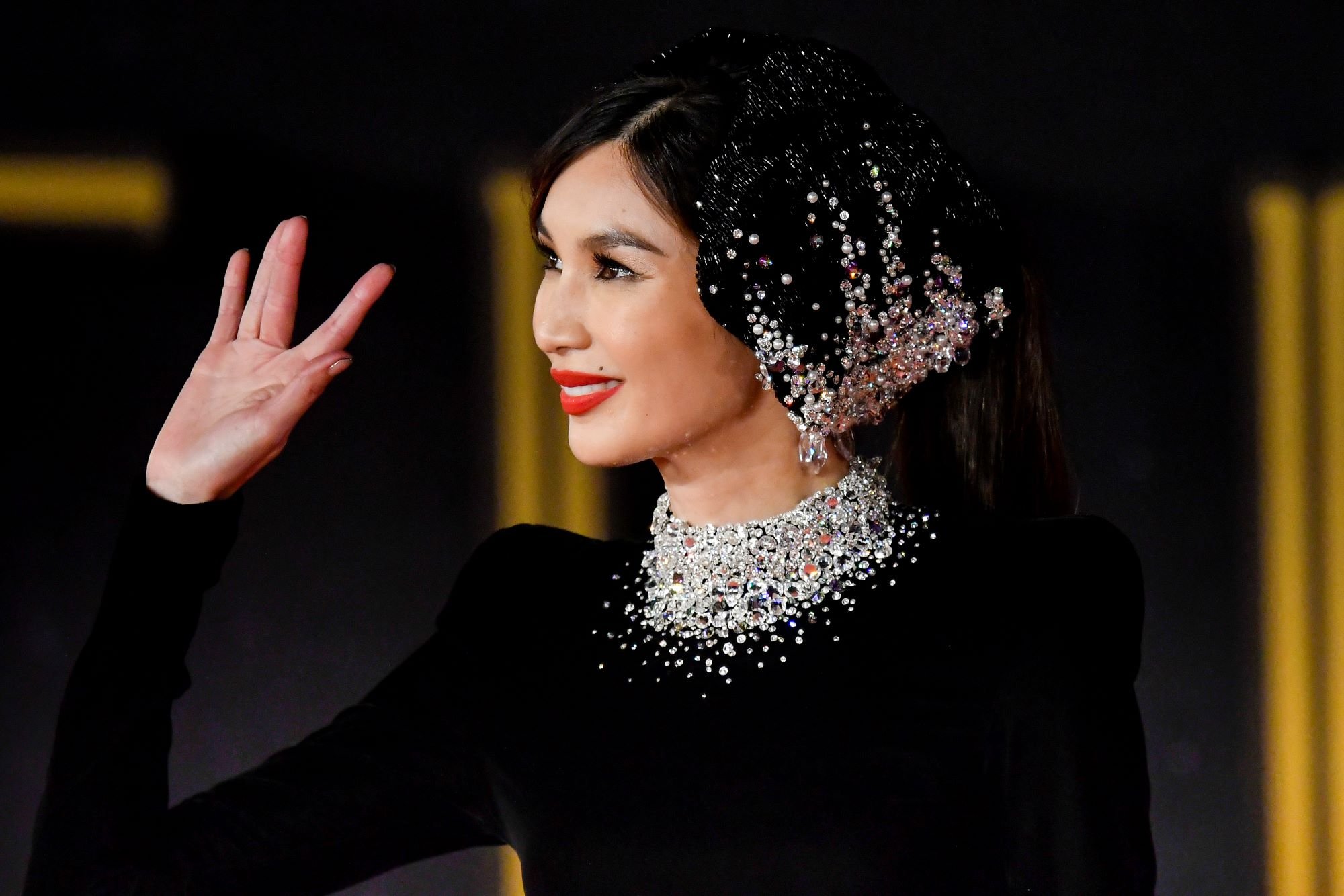 Marvel's 'Eternals' star Gemma Chan waves and wears a long-sleeved black dress with a diamond-studded collar and a black headpiece with numerous gems on it.