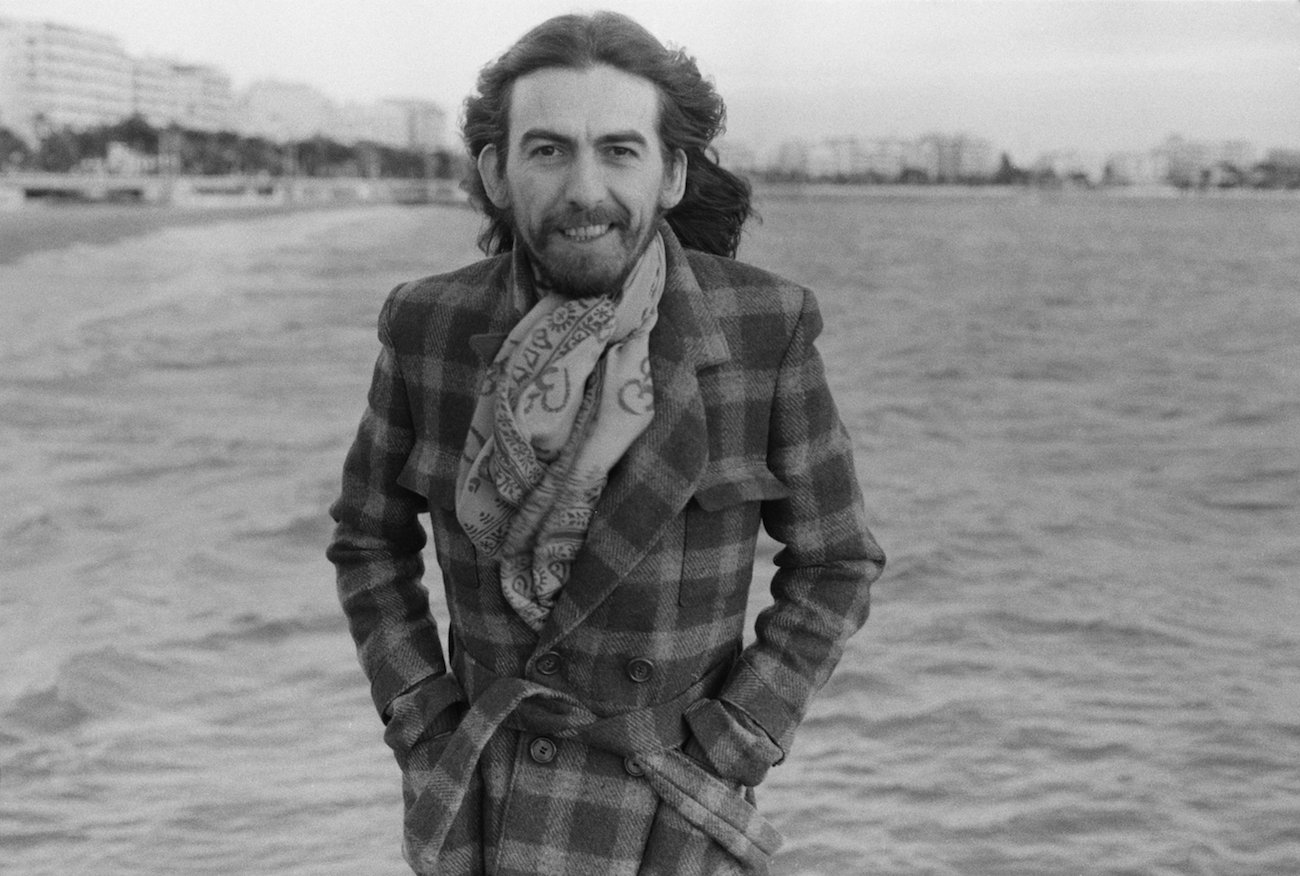 George Harrison wearing a jacket and scarf on the beach in Cannes, France, 1976.
