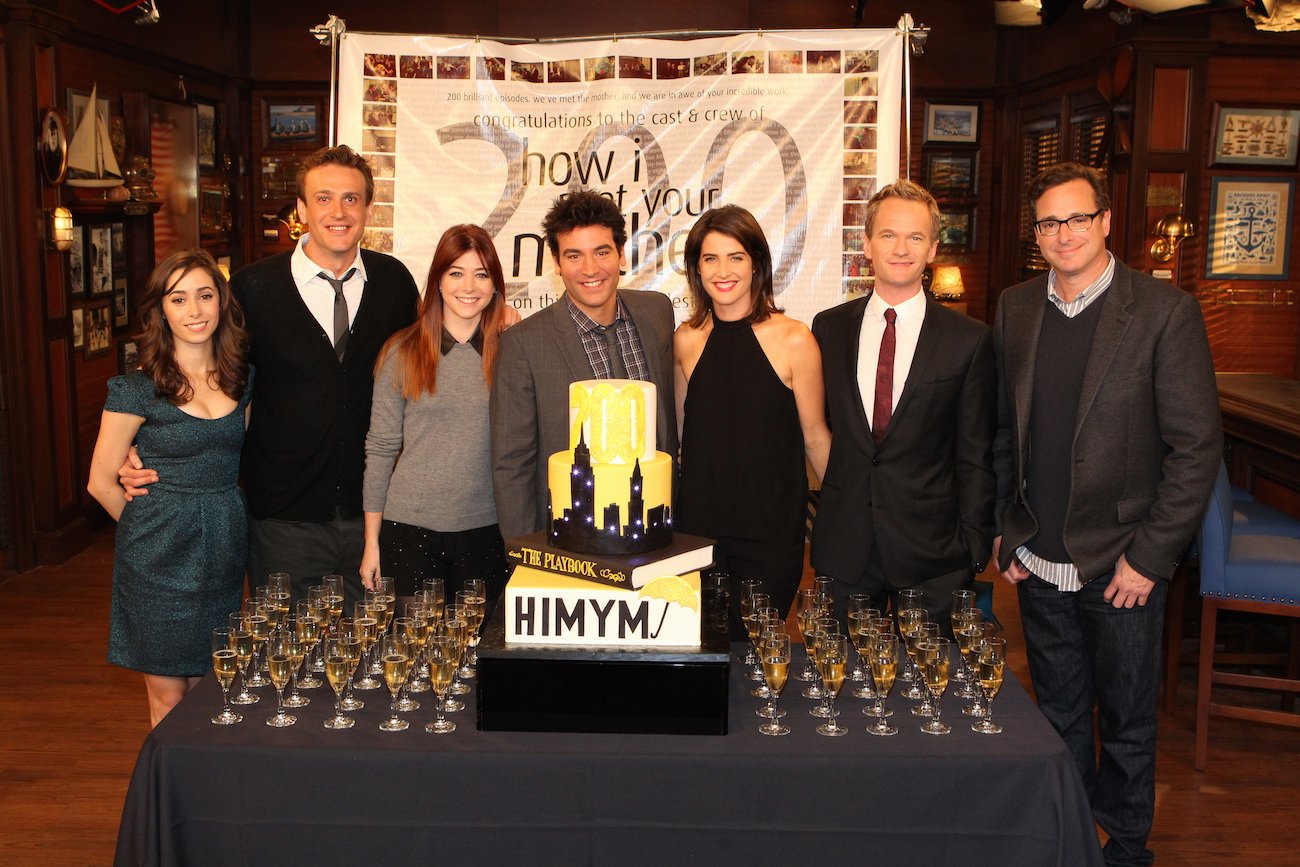 Bob Saget with other cast members of 'How I Met Your Mother' standing behind a table with a cake and wine glasses