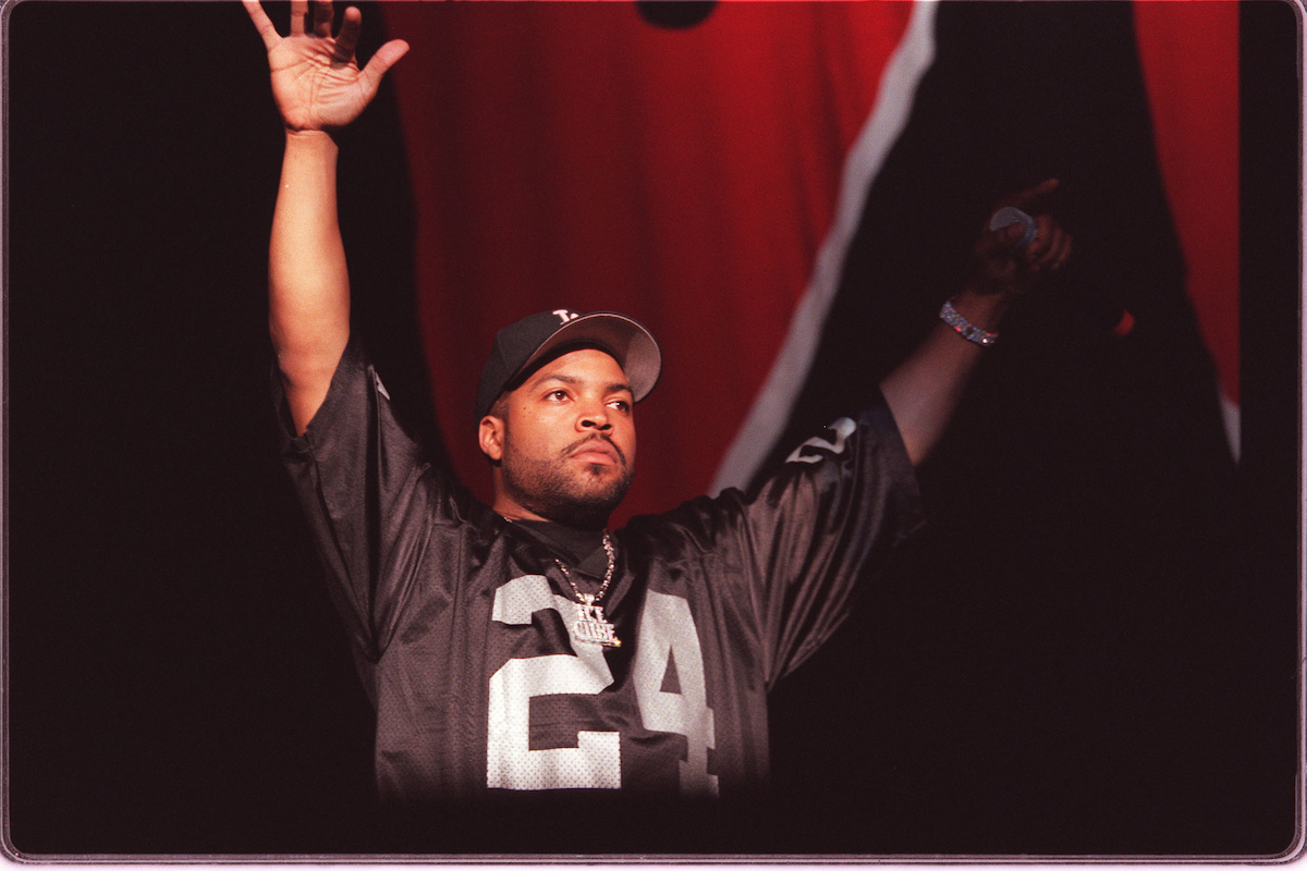 Ice Cube wears a football jersey and cap as he puts his arms up while on stage