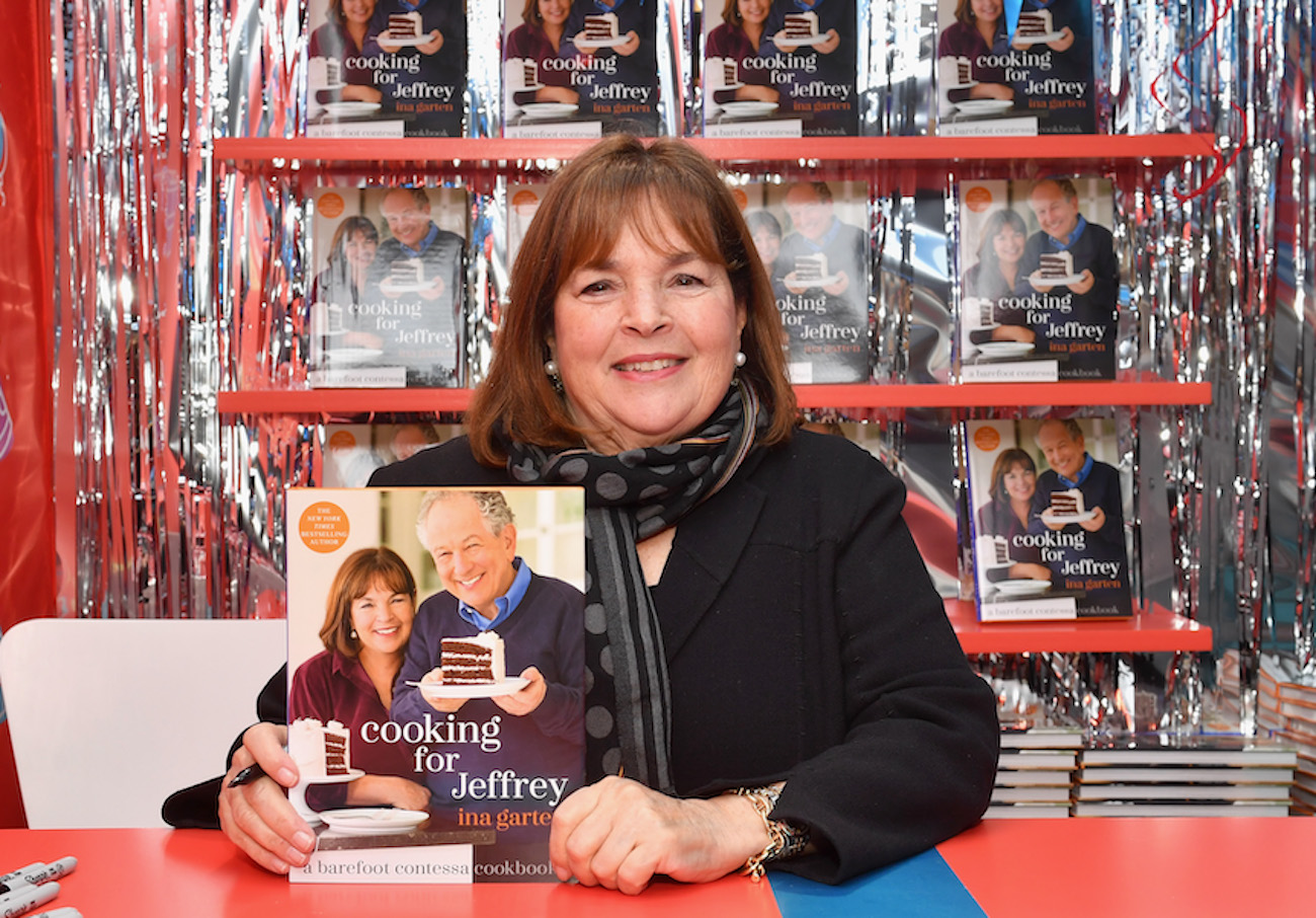 Ina Garten smiles as she sits at a table holding a copy of 