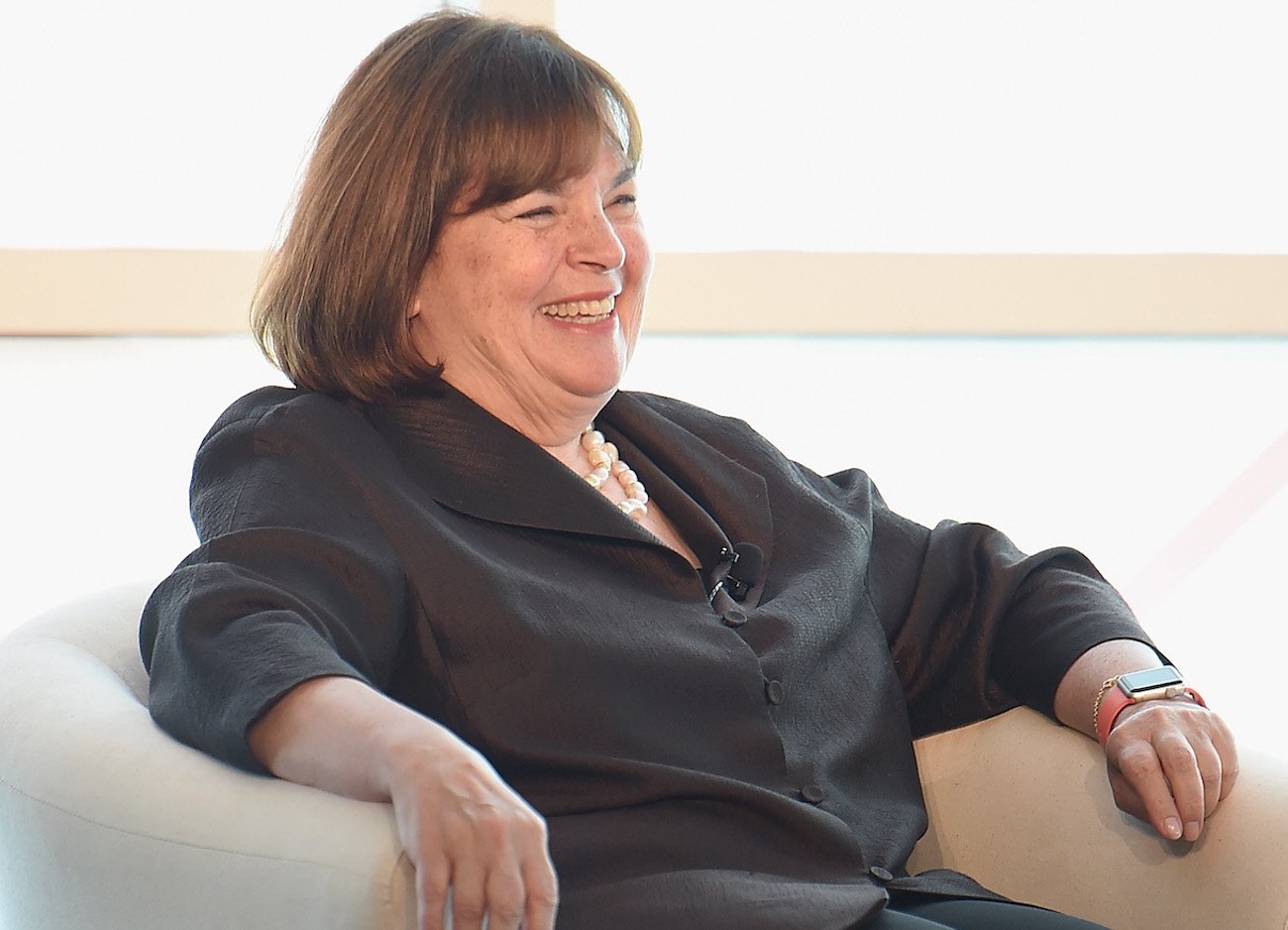 Ina Garten smiles as she sits in a chair wearing a black button down shirt