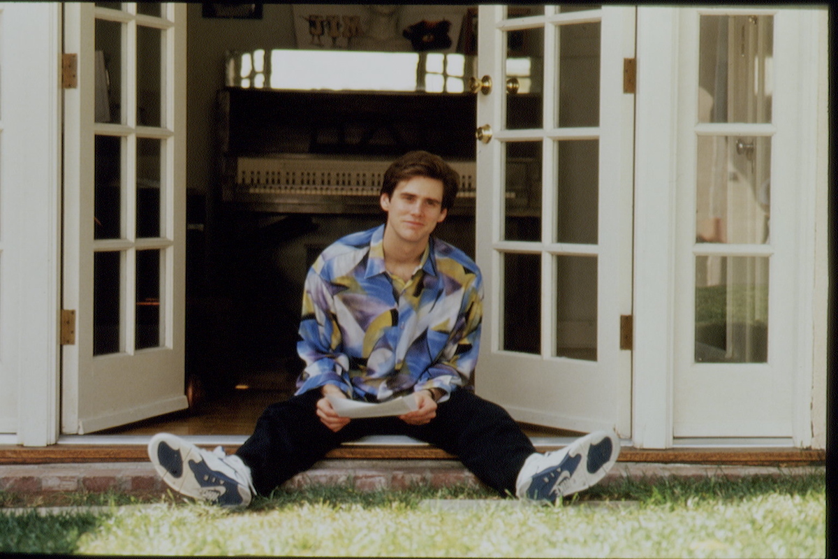 Jim Carrey sits in front of two open double doors looking off into the distance