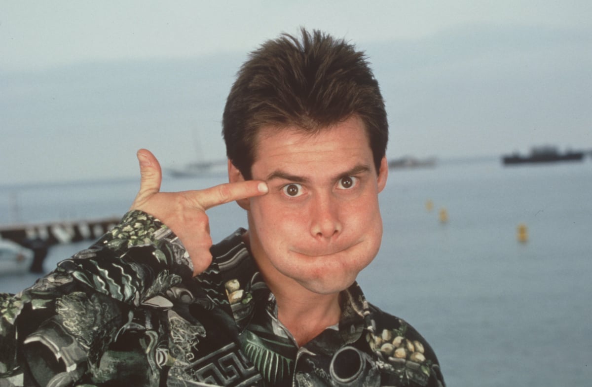Jim Carrey makes a funny face and points to his temple in a Hawaiian shirt