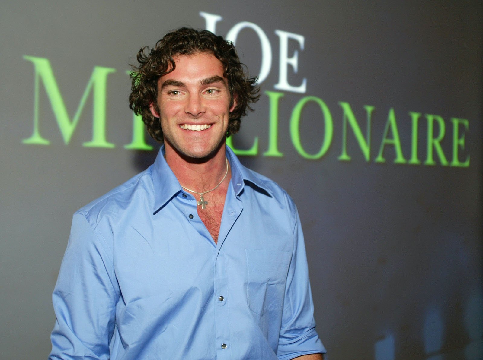 'Joe Millionaire' star Evan Marriott smiles wearing a blue shirt while standing in front of a backdrop with the show's title.