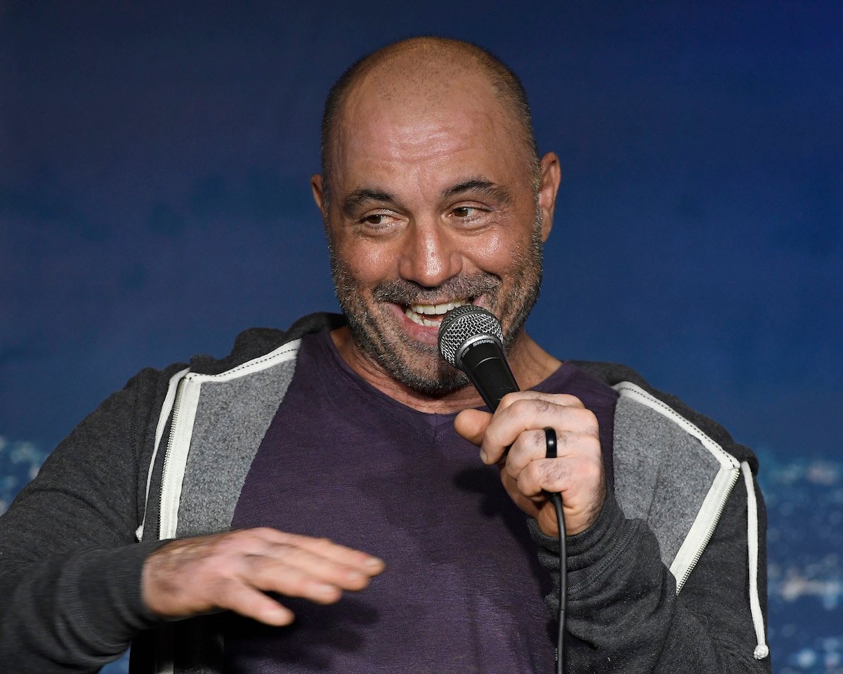 Joe Rogan holds a microphone and smiles.