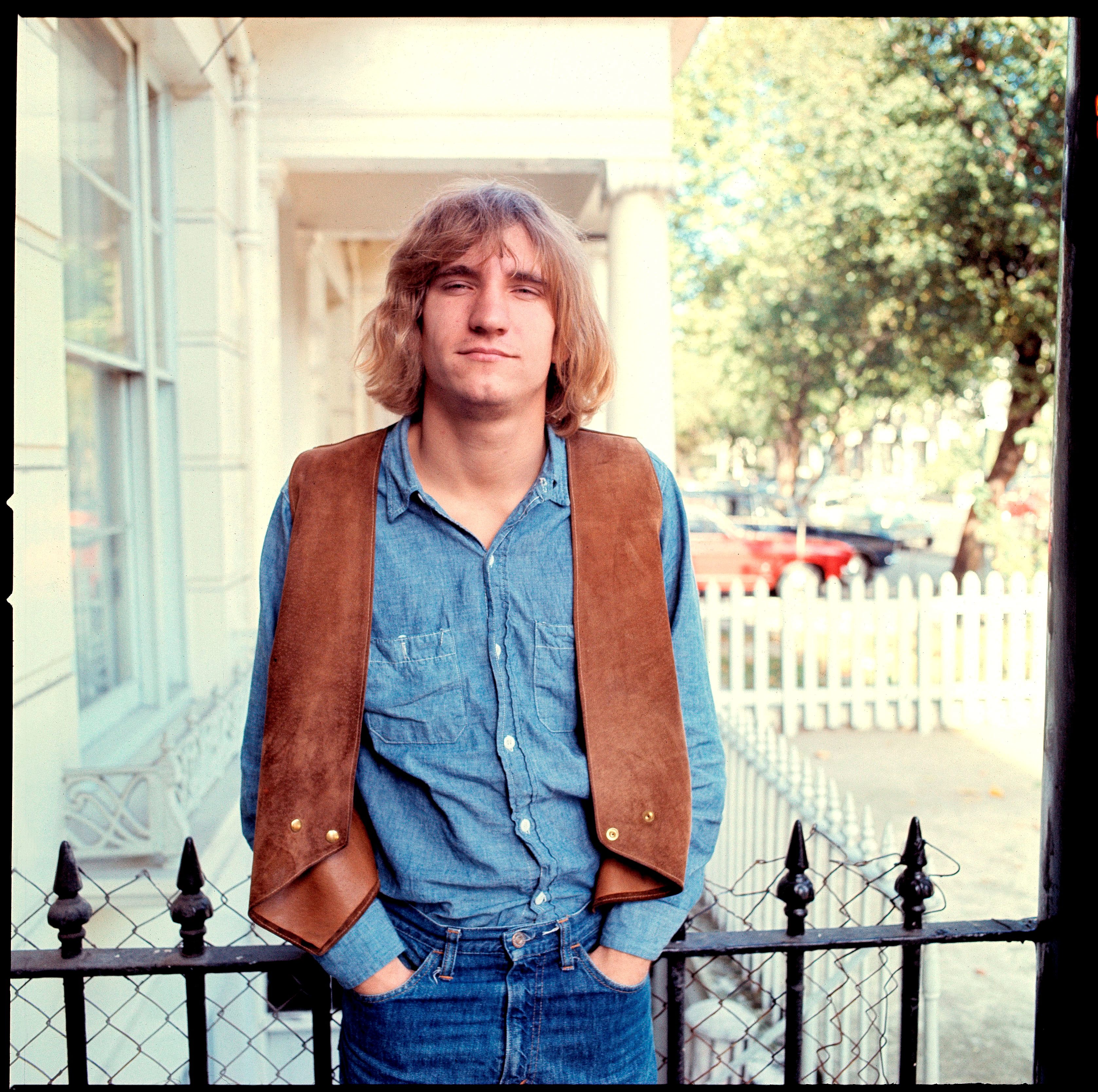 Joe Walsh wears a denim outfit and a blue vest. He stands on a porch.