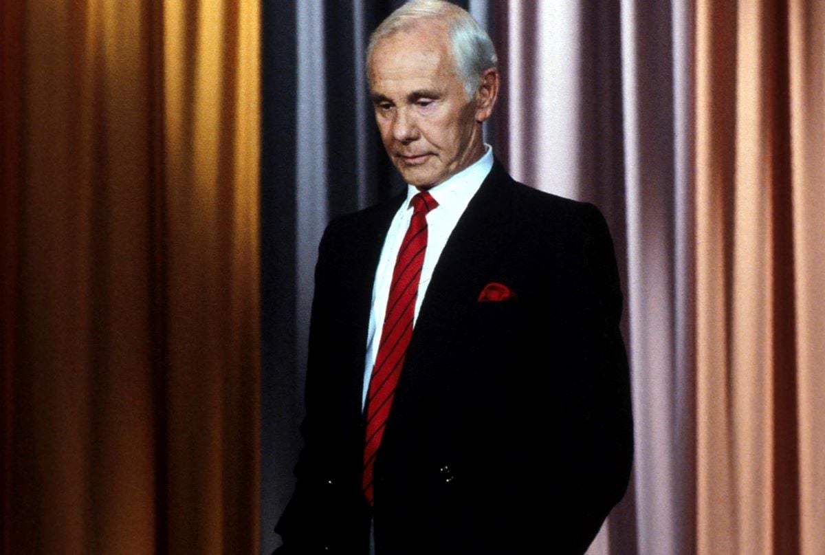 Johnny Carson in a black jacket and red tie, hands in pockets and looking down