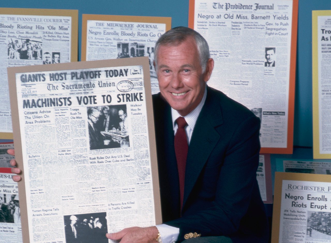 Johnny Carson smiles while he holds up a newspaper with a headline that reads 'Machinists Vote to Strike'