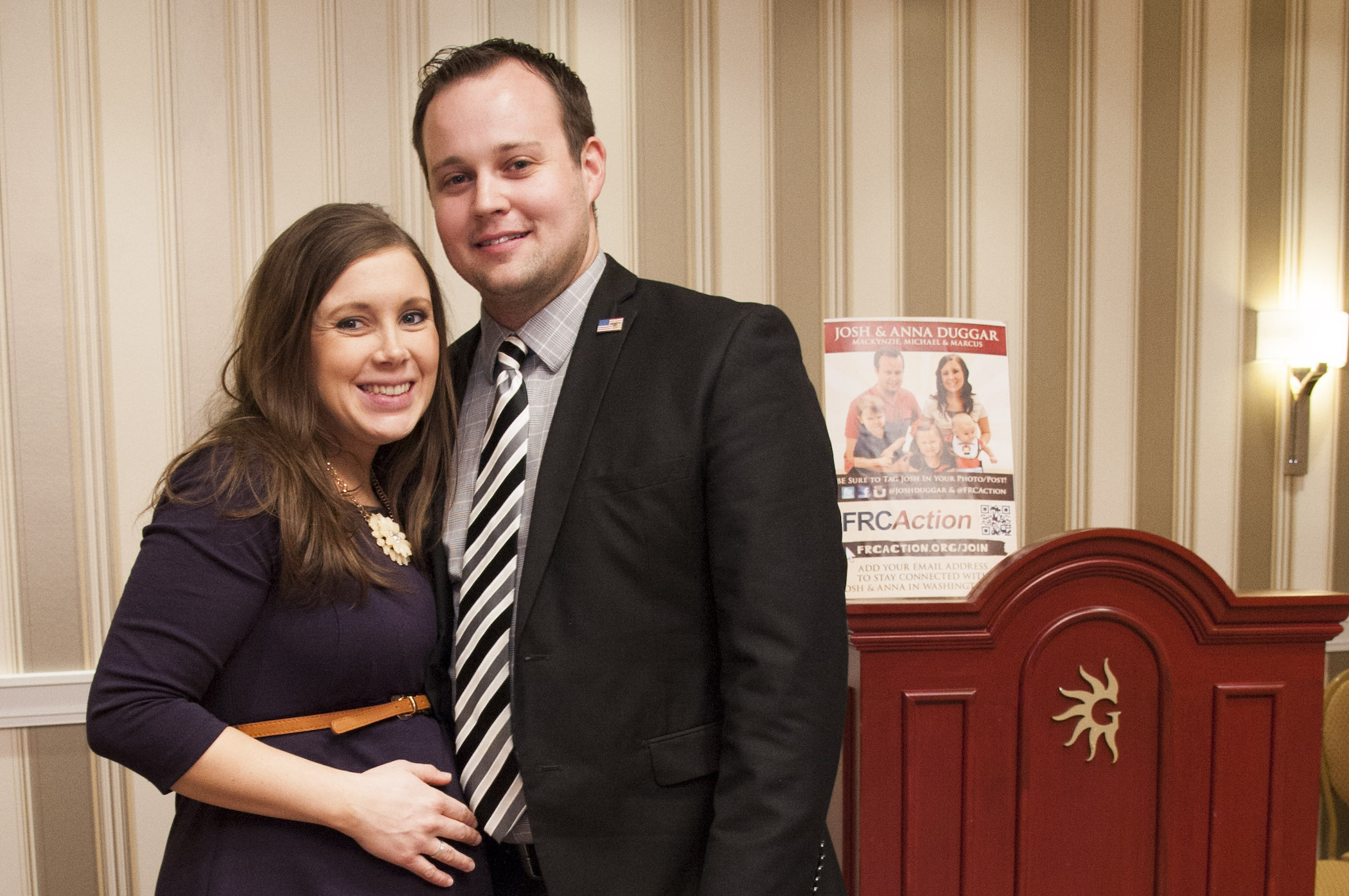 Anna Duggar and Josh Duggar standing together and smiling at an event. Anna is pregnant and holding her belly with one hand