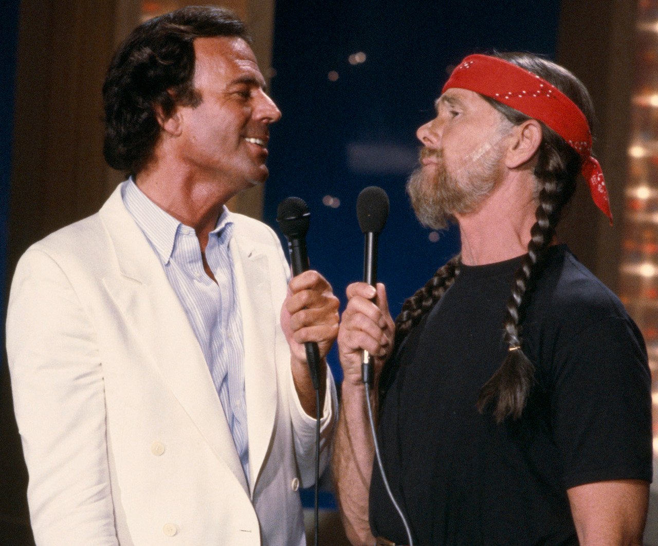 Julio Iglesias in a white suite, holding a microphone and looking at Johnny Carson, who is dressed as Willie Nelson