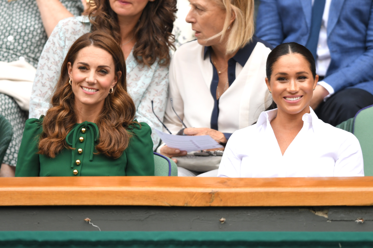 Kate Middleton and Meghan Markle smiling and sitting side by side. Kate is wearing a green dress and Meghan is wearing a white shirt.
