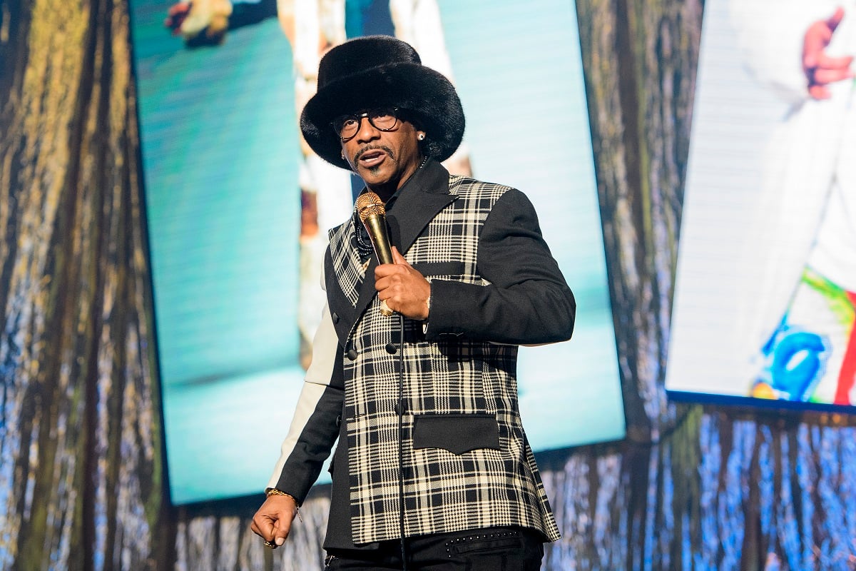 Katt Williams wearing a grey suit while holding a gold microphone.