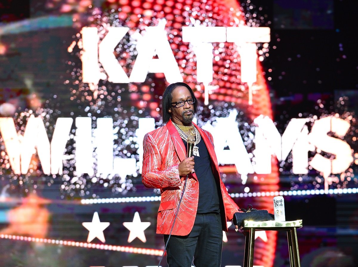 Katt Williams holding a microphone and wearing a red jacket.