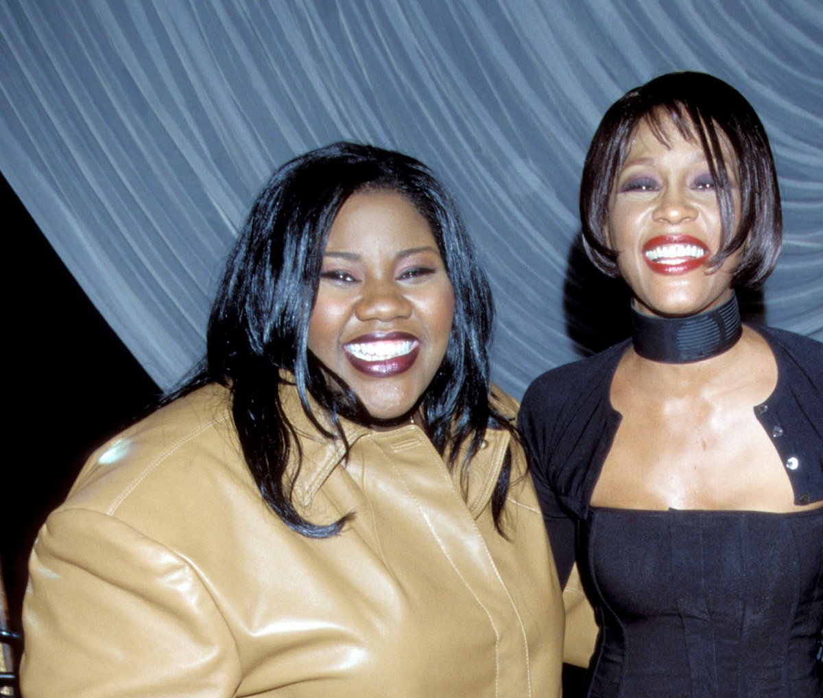Kelly Price wearing a tan leather jacket and Whitney Houston wearing a black dress pose for photo.