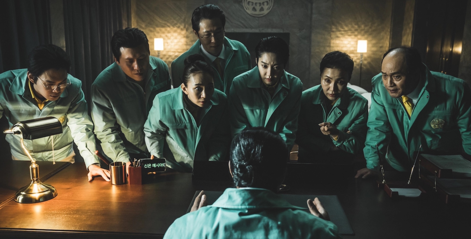Kim Mi-soo in 'Hellbound' episode 6 wearing green jacket leaning over desk.