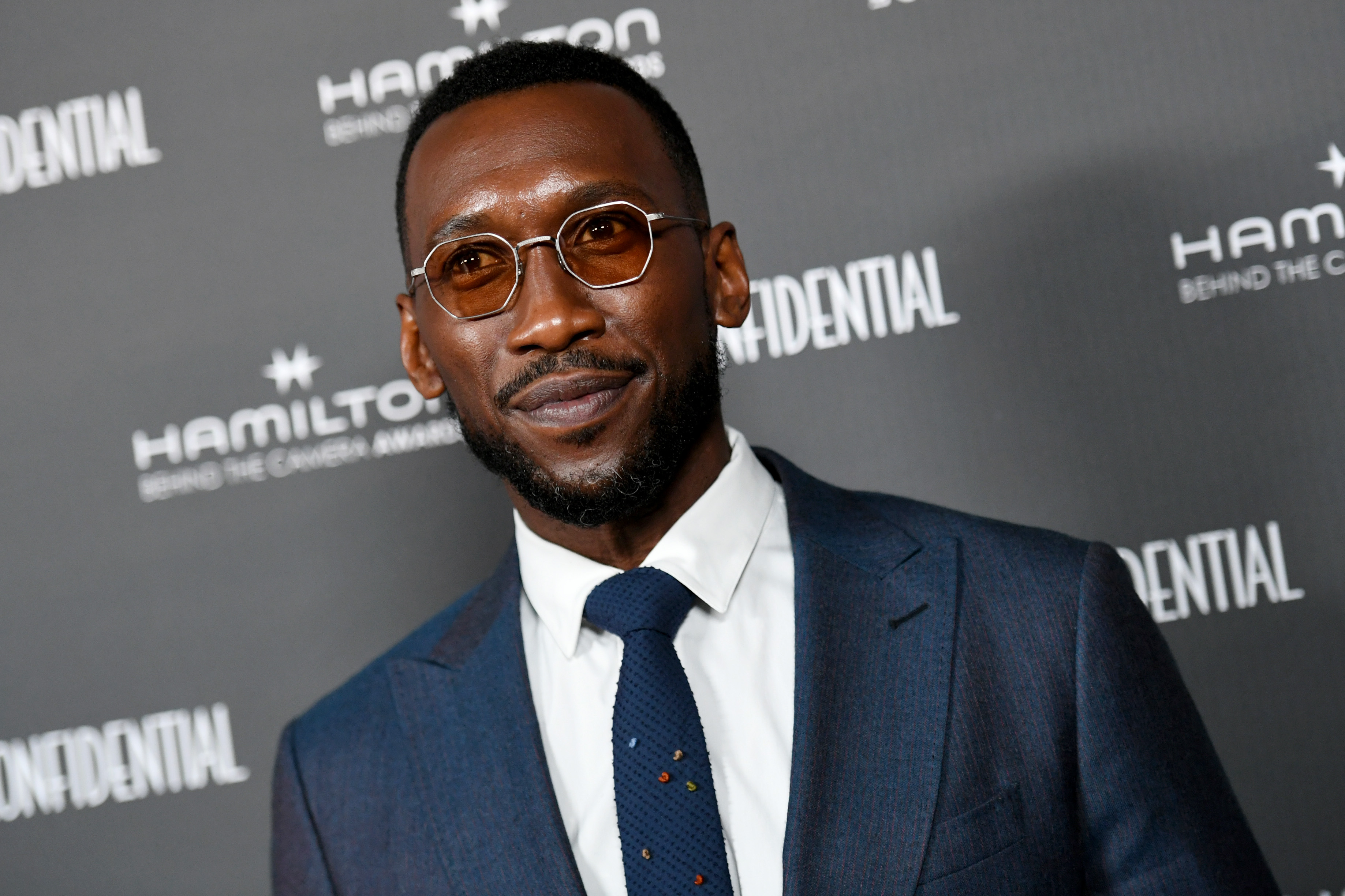 ‘Blade’ actor Mahershala Ali, heard in Marvel’s ‘Eternals’ post-credits scene, wears a dark blue suit jacket over a white button-up shirt and dark blue tie and wears glasses.

