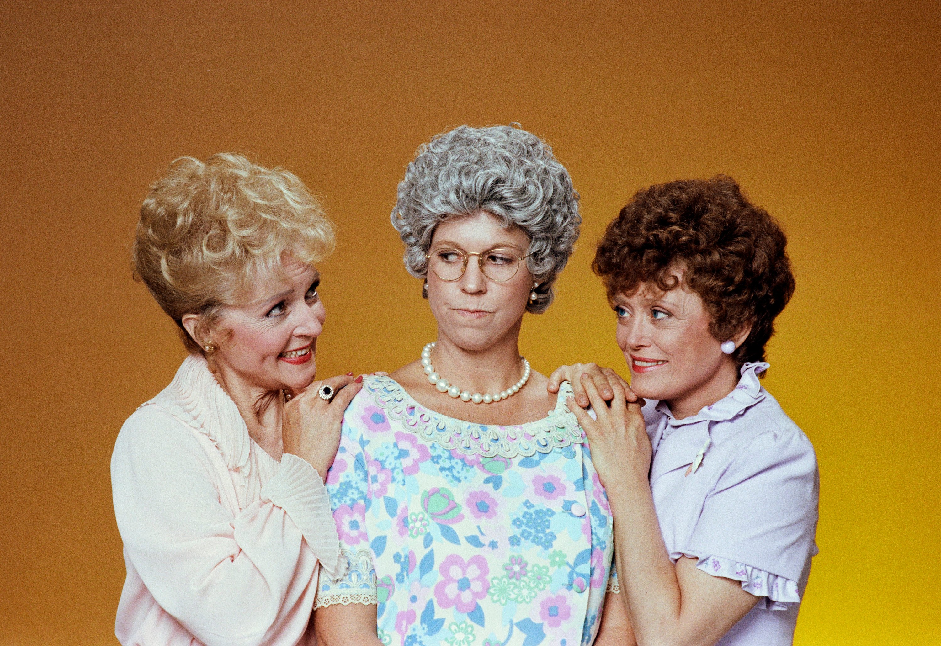 'Mama's Family' actors Betty White, Vicki Lawrence, and Rue McClanahan dressed as their characters and standing in front of a yellow backdrop.