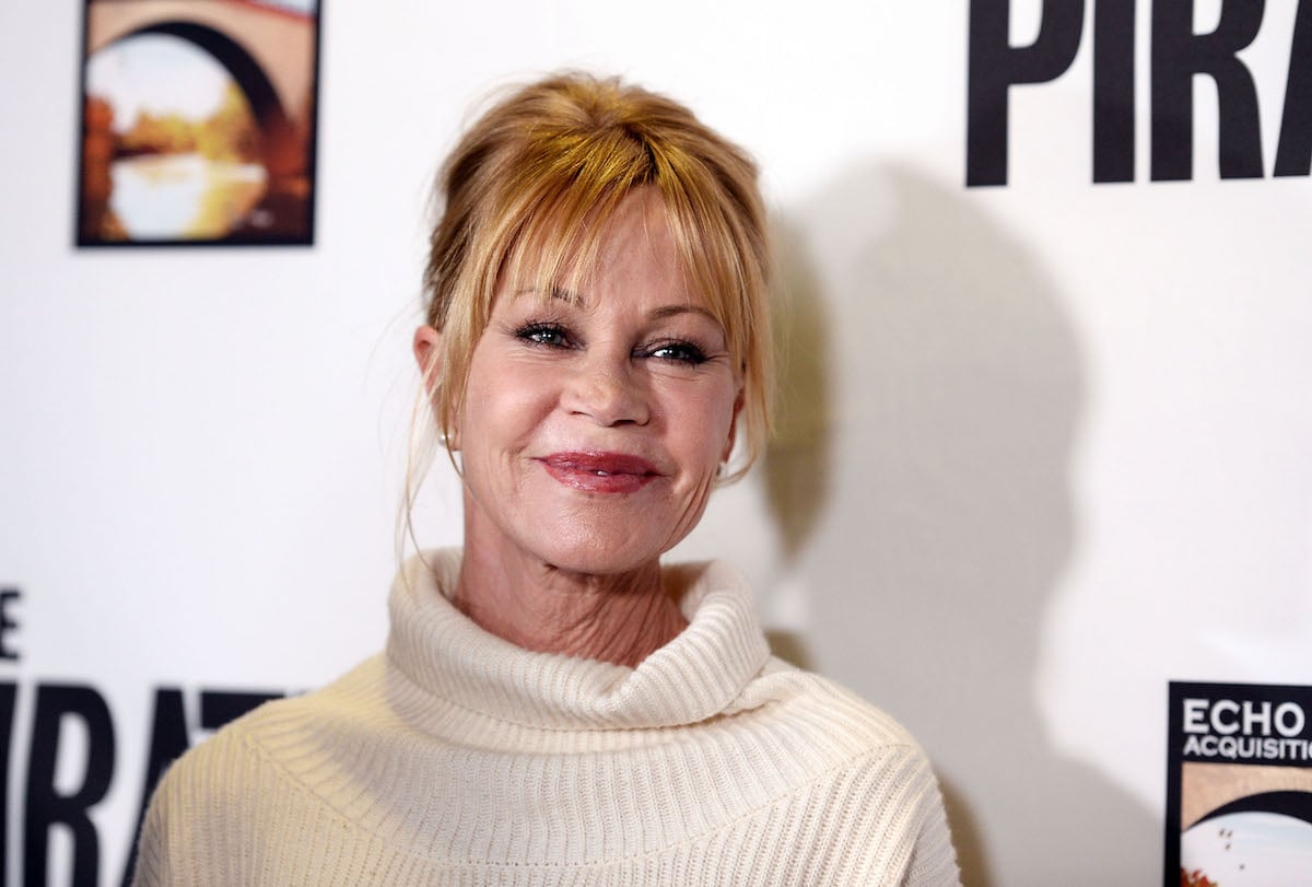 A closeup of Melanie Griffith in a white turtleneck sweater and her hair up.