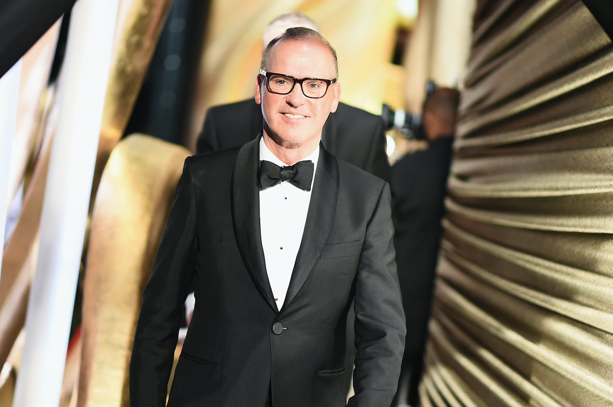 Michael Keaton wears a black suit and glasses while smiling on stage