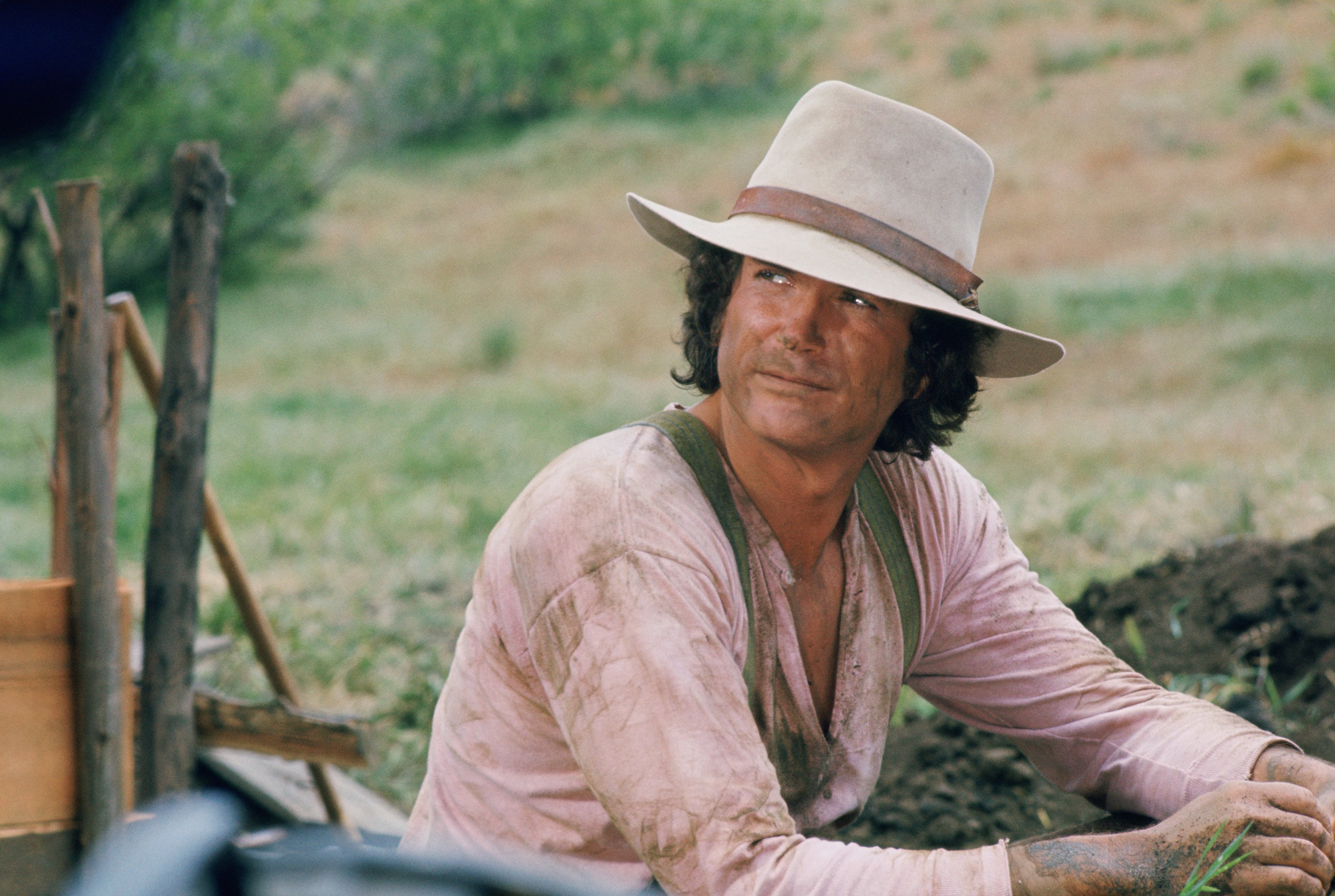 Michael Landon wears a hat and looks up while sitting on the farm on the set of 'Little House on the Prairie.'